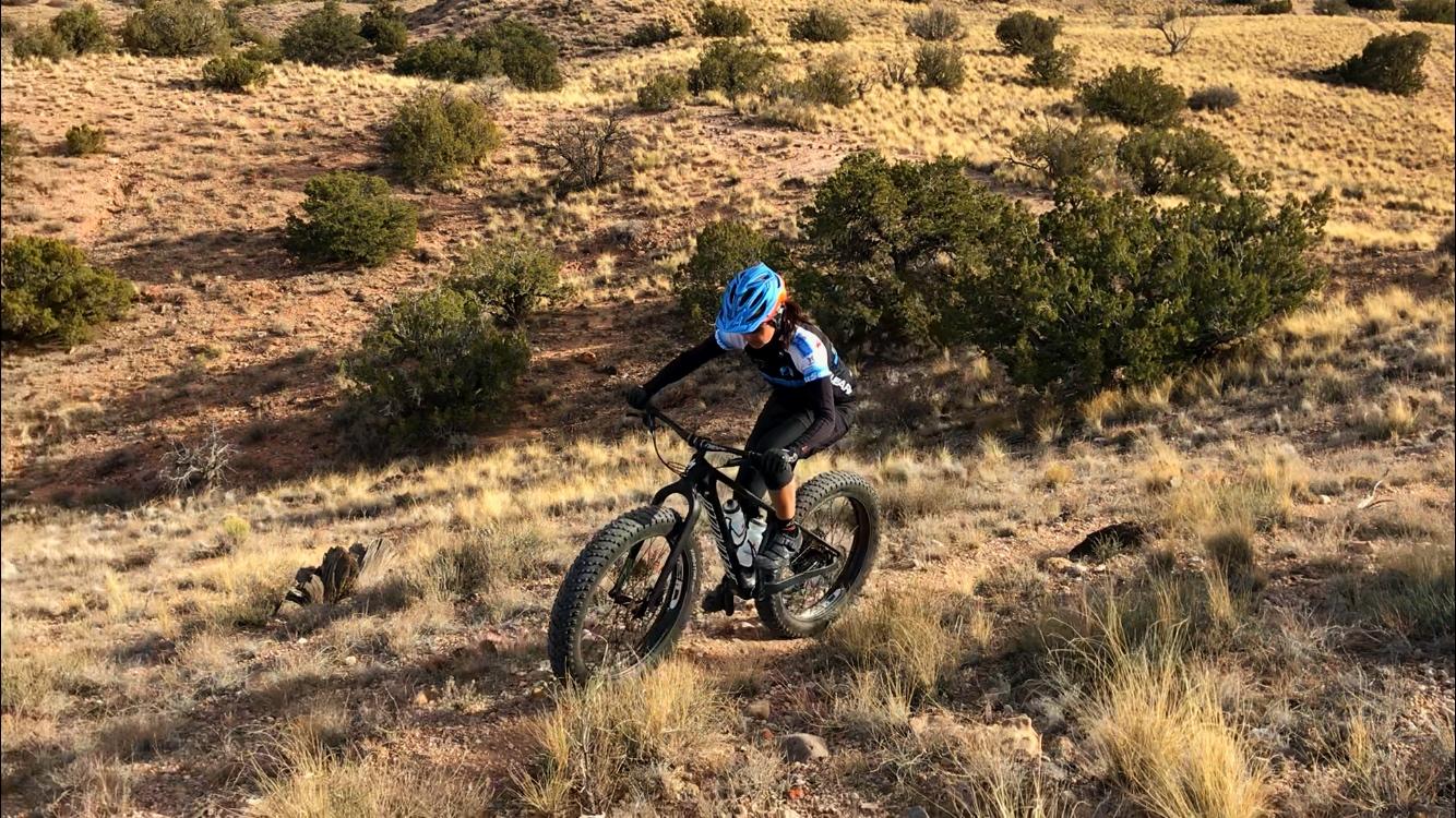 A person riding a fat-tire bicycle on a rugged, hilly terrain with sparse vegetation and dry grass. The cyclist is wearing a blue helmet and a sports jersey, navigating uphill amidst a backdrop of trees and rocky ground. Mariposa Fat Bike Trails mountain bike trail.