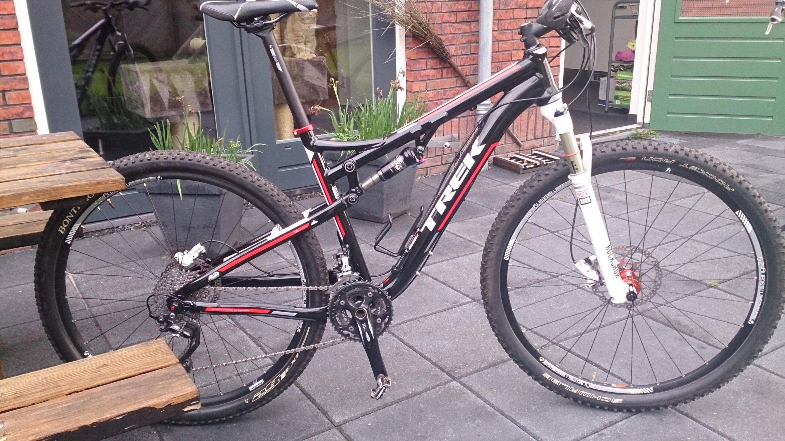 Trek Superfly AL Elite: A black and red Trek mountain bike is parked beside a wooden picnic table on a paved patio. The bike features thick tires and a dual suspension system, showcasing its suitability for off-road riding. In the background, there are green plants and a glimpse of a house with a green door.