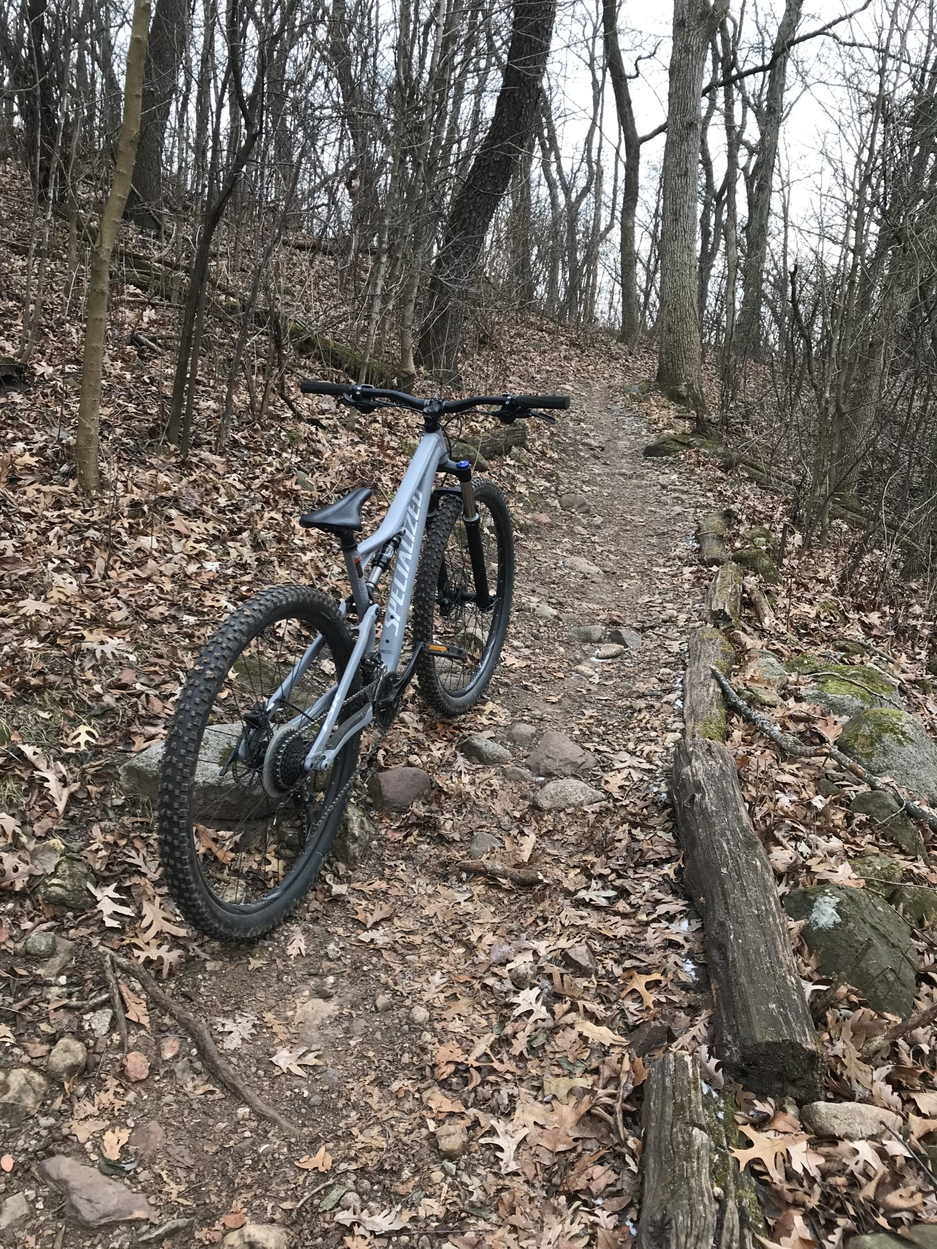 A mountain bike is parked on a winding dirt trail surrounded by bare trees and autumn leaves. The path is rocky and lined with logs, indicating a natural outdoor setting ideal for biking. Kettle Moraine John Muir + Emma Carlin mountain bike trail.