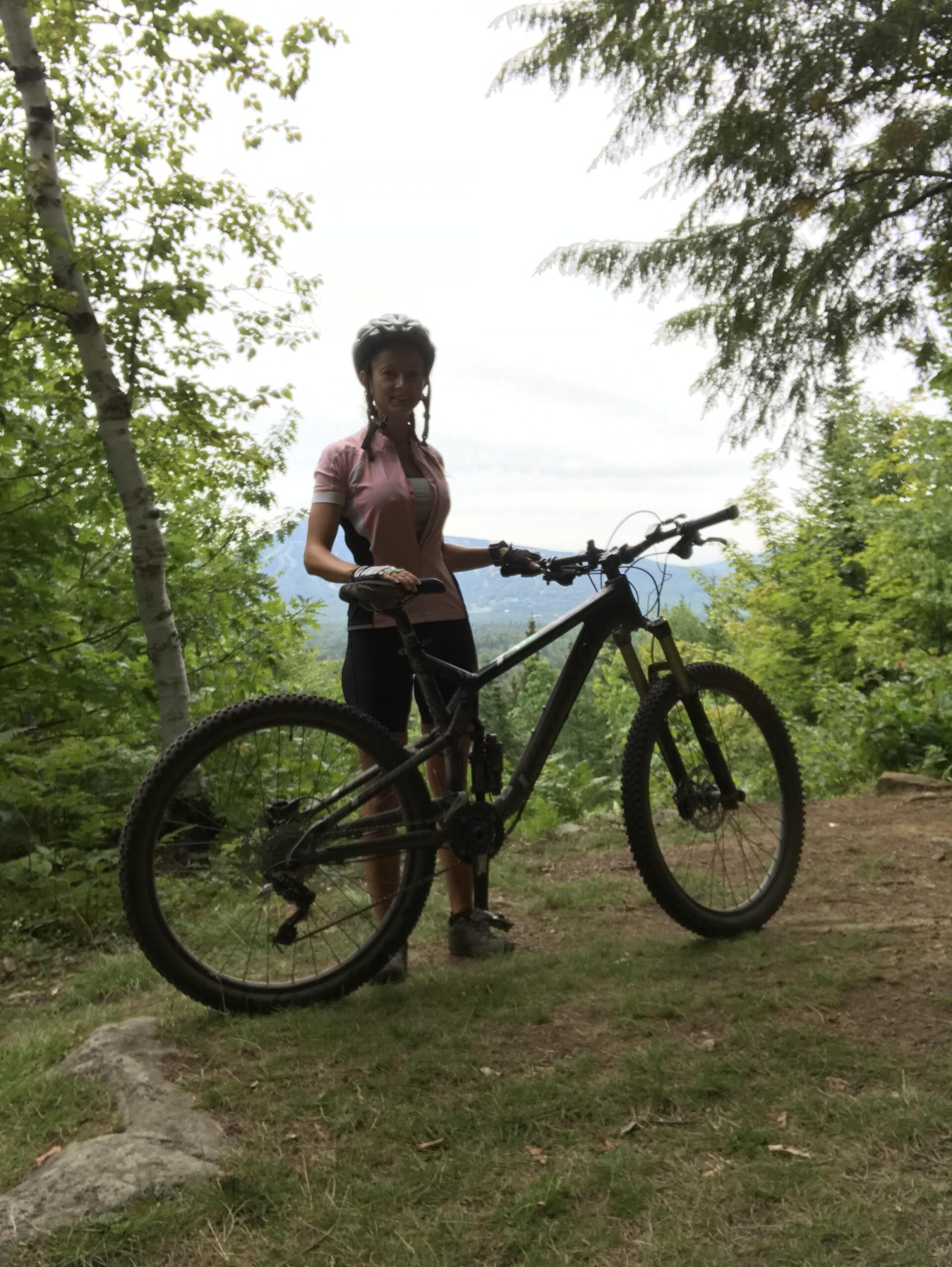 Cannondale Trigger Carbon 2 27.5: A person wearing a helmet and cycling attire stands next to a mountain bike on a trail surrounded by greenery, with a scenic view in the background.