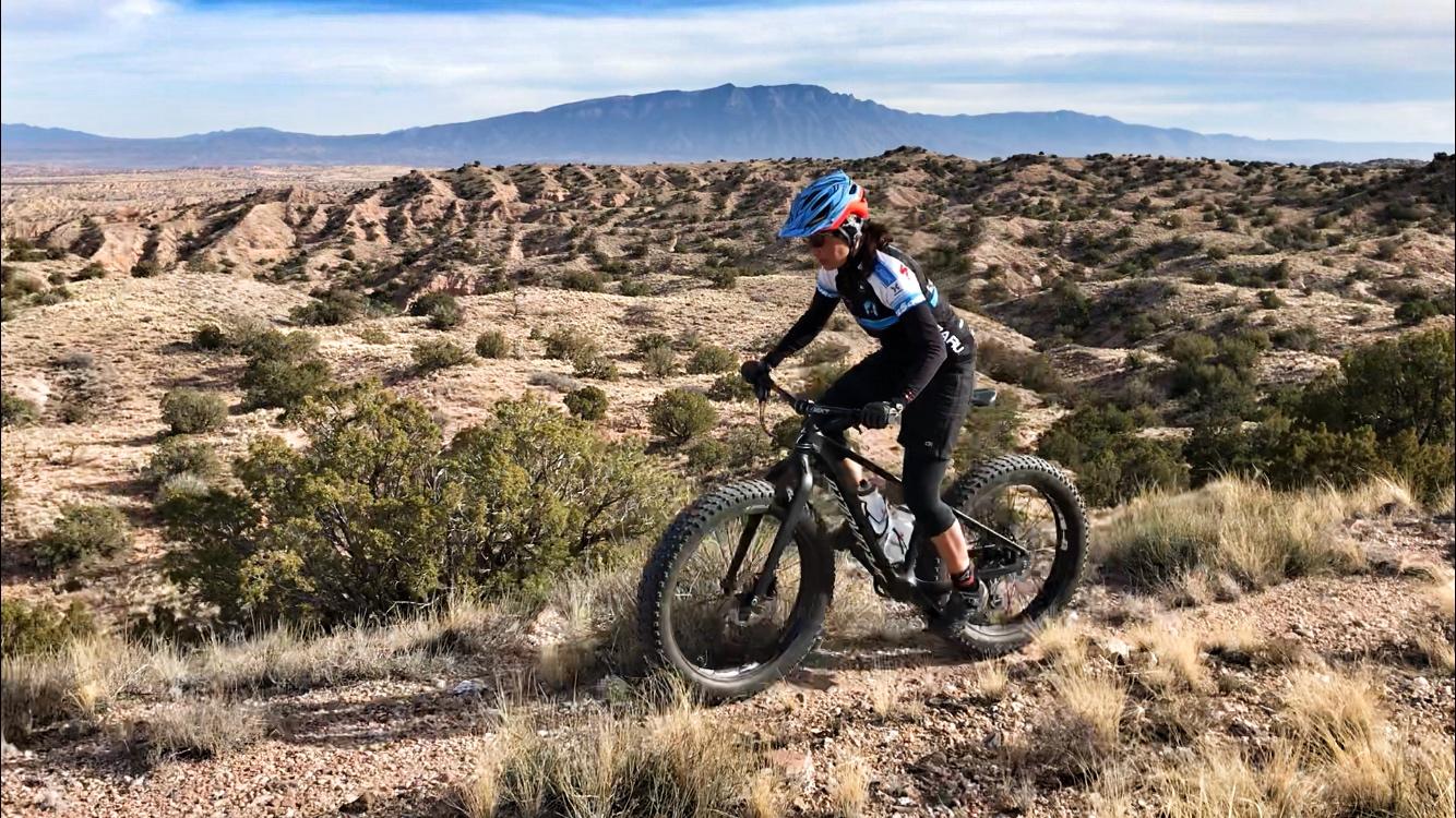 A person riding a mountain bike on a rugged trail, surrounded by picturesque desert landscapes and distant mountains under a blue sky. The rider is wearing a helmet and athletic gear, showcasing an adventurous spirit in a natural outdoor setting. Mariposa Fat Bike Trails mountain bike trail.