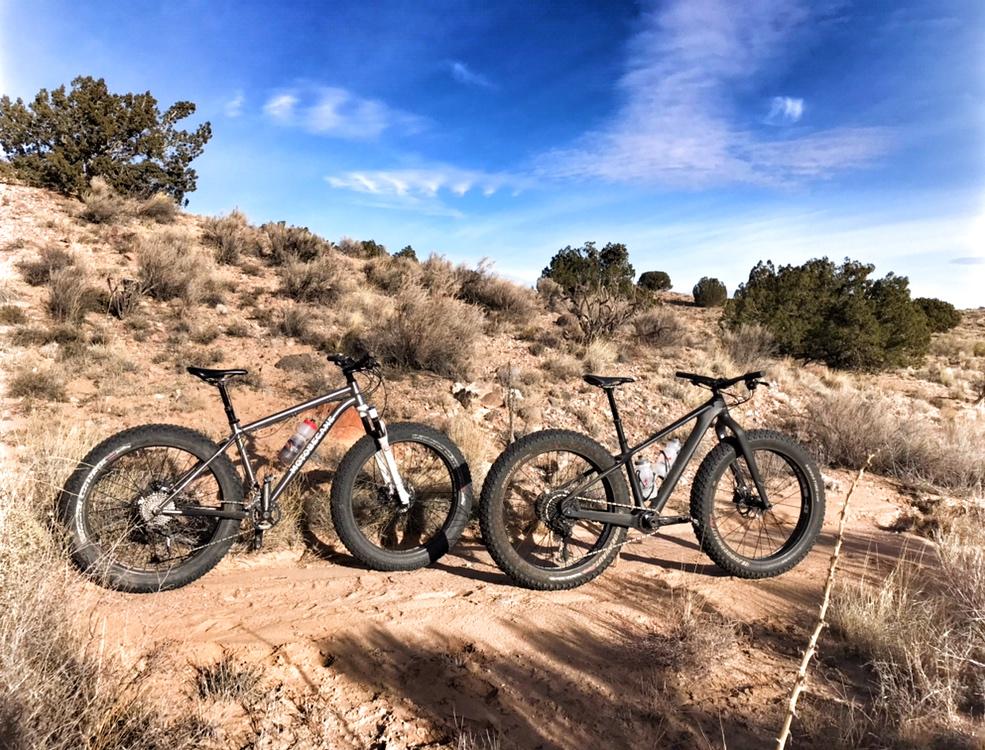 Two fat tire mountain bikes parked on a sandy trail surrounded by sparse vegetation and rocky terrain, under a blue sky with scattered clouds. Mariposa Fat Bike Trails mountain bike trail.