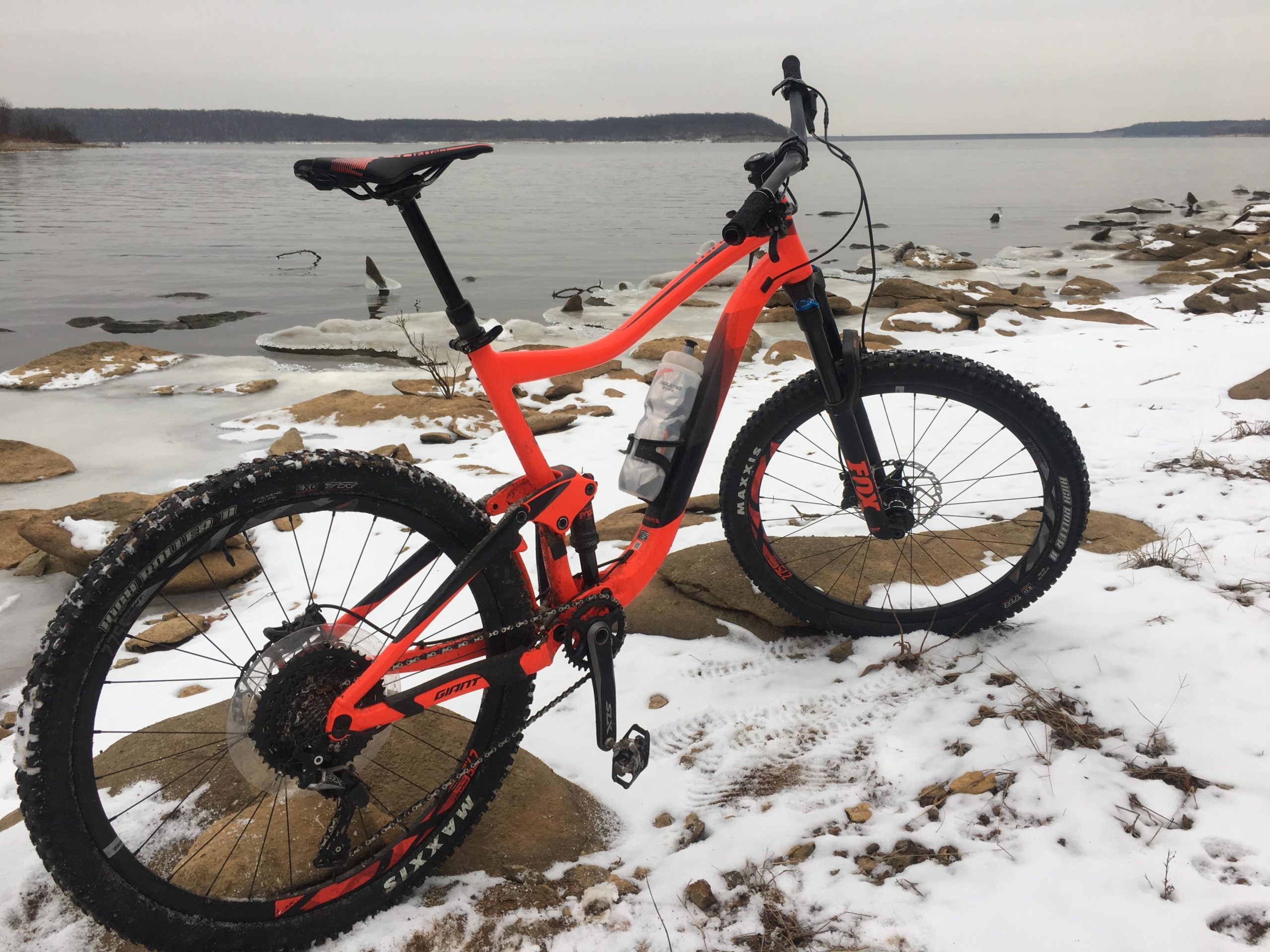 A bright orange mountain bike parked on rocky terrain near a frozen lake, surrounded by patches of snow. The bike features thick tires and a water bottle attached to the frame, with a cloudy sky in the background. Perry Lake Bike Trail mountain bike trail.