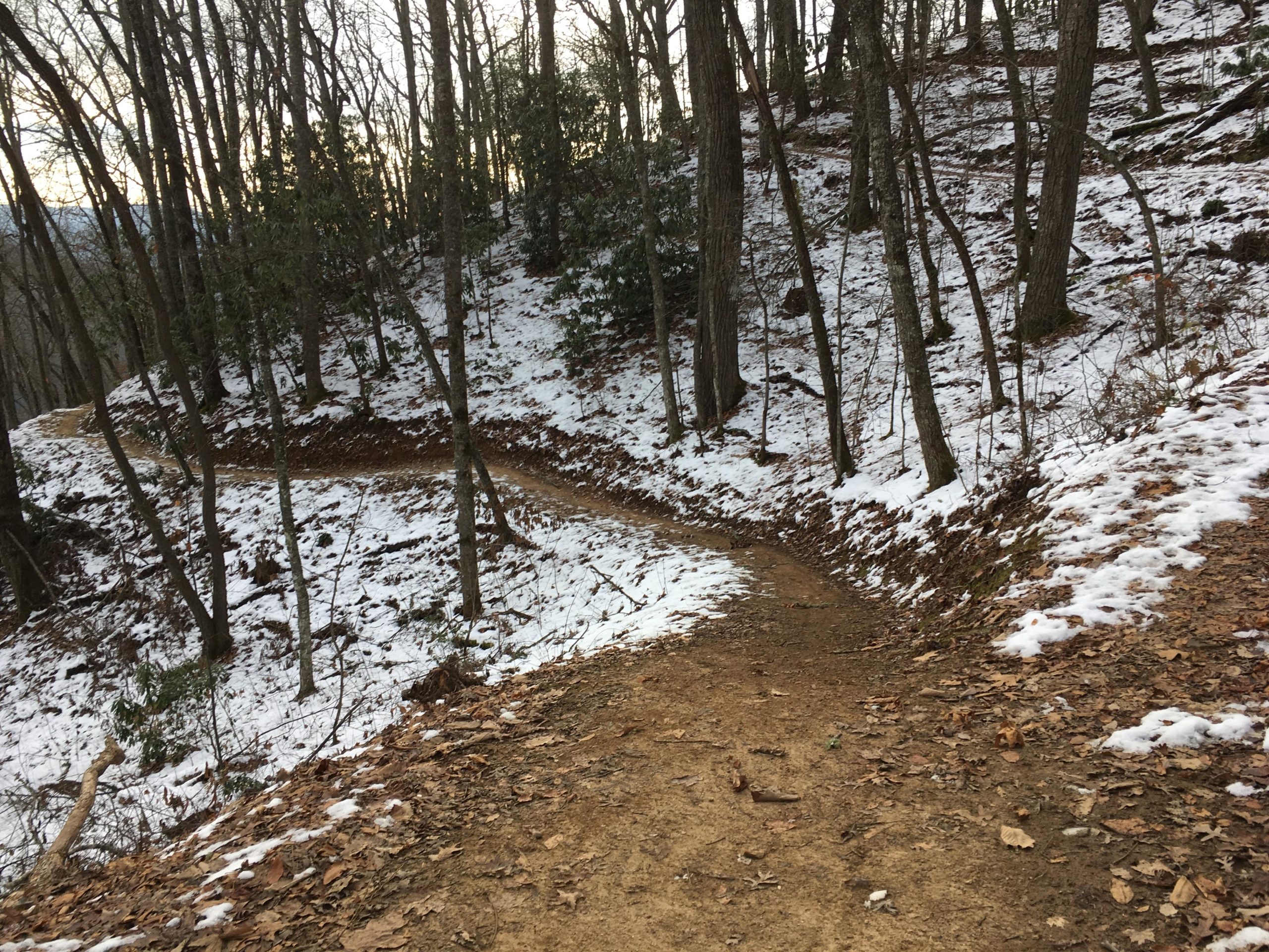 A winding dirt trail surrounded by bare trees on a snowy hillside, with patches of snow and fallen leaves visible along the path. Fire Mountain Trail System mountain bike trail.