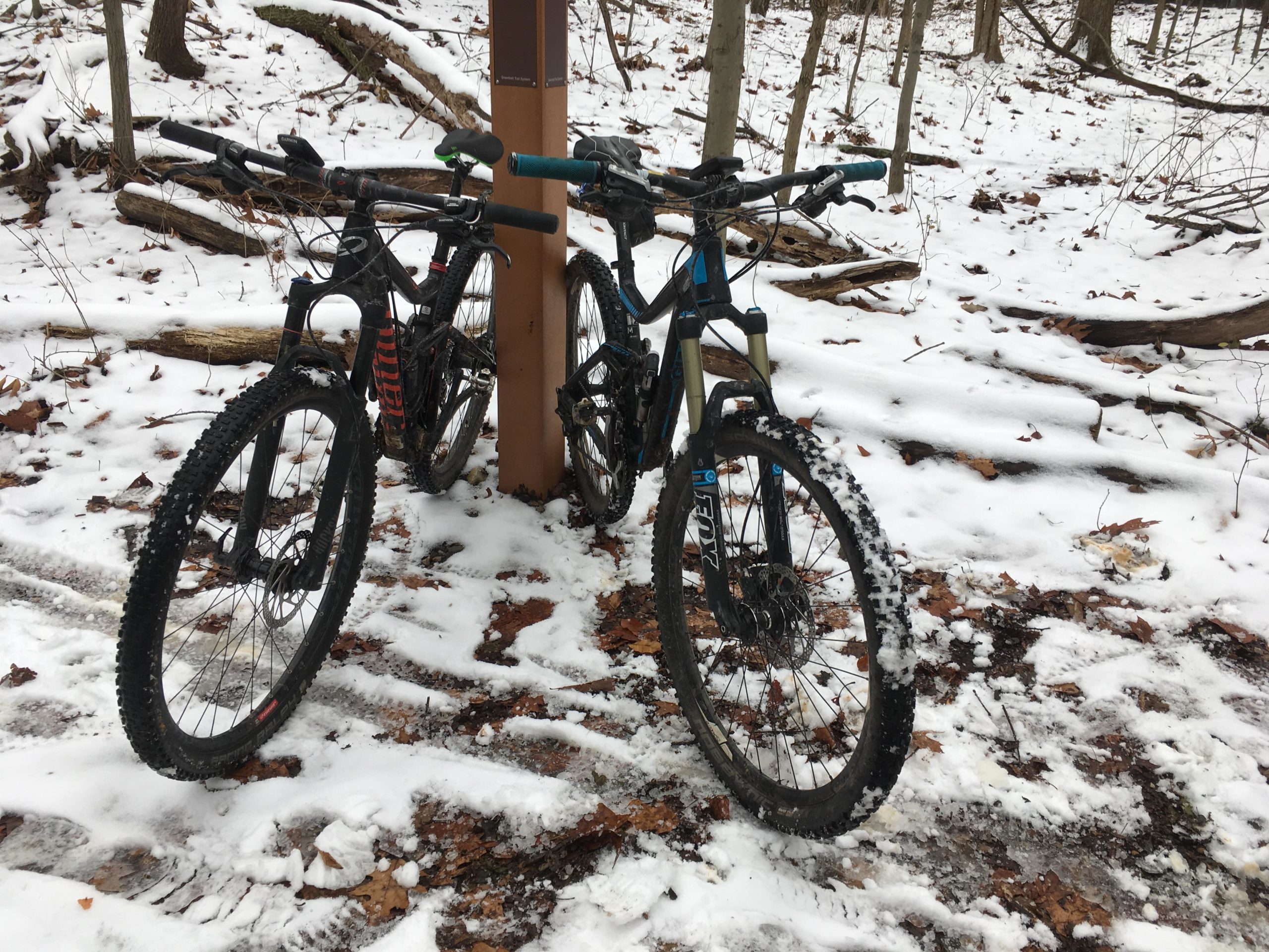 Two mountain bikes are resting against a trail marker in a snowy forest. The ground is covered in snow with some visible leaves scattered around, and bare trees can be seen in the background. Richmond Avenue and Forest Hill road mountain bike trail.