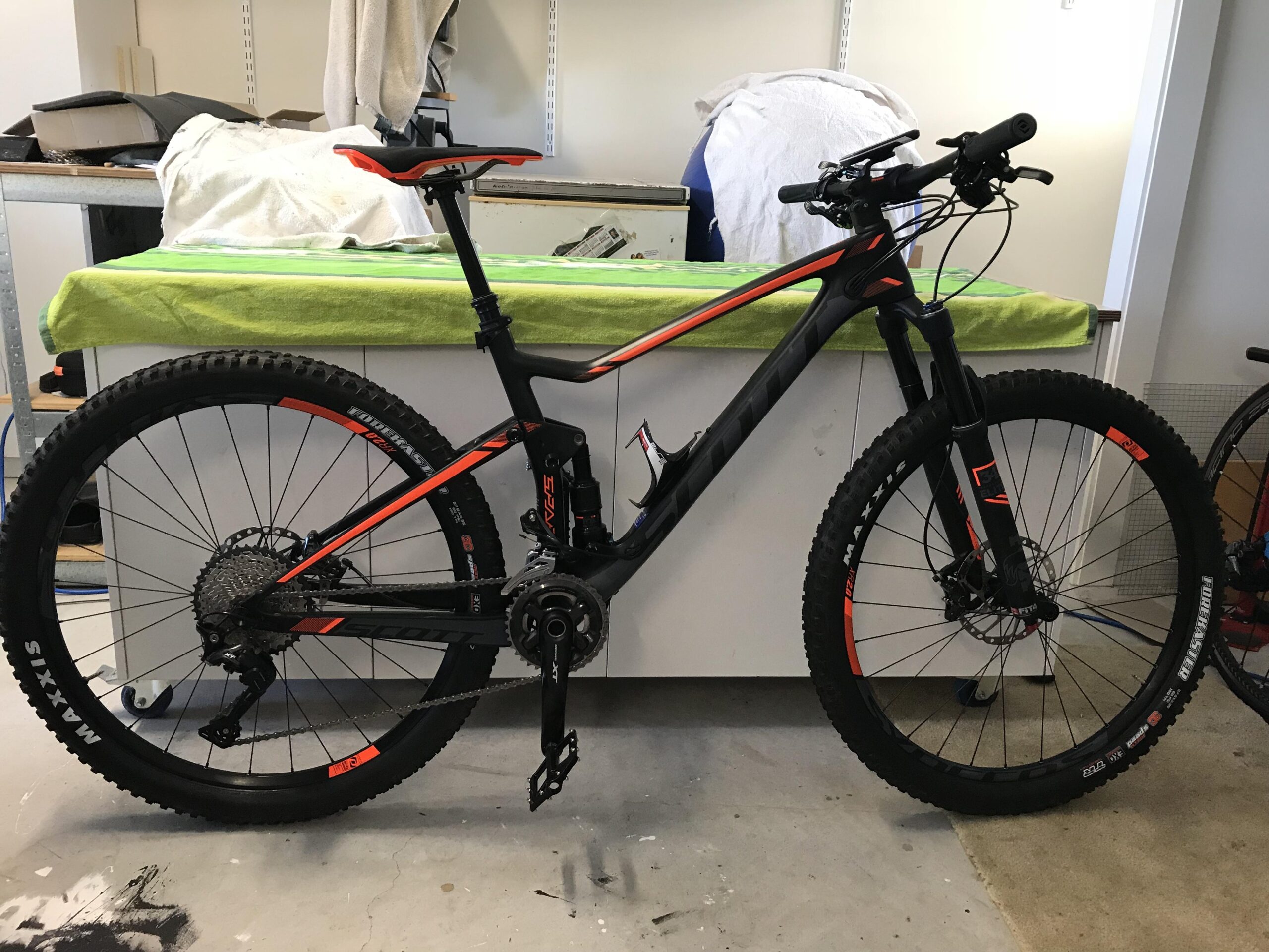 Scott Spark 710: A black and orange mountain bike is positioned in a workshop setting, with a workbench in the background covered in a green towel. The bike features a suspension frame, thick tires, and a visible gear system.