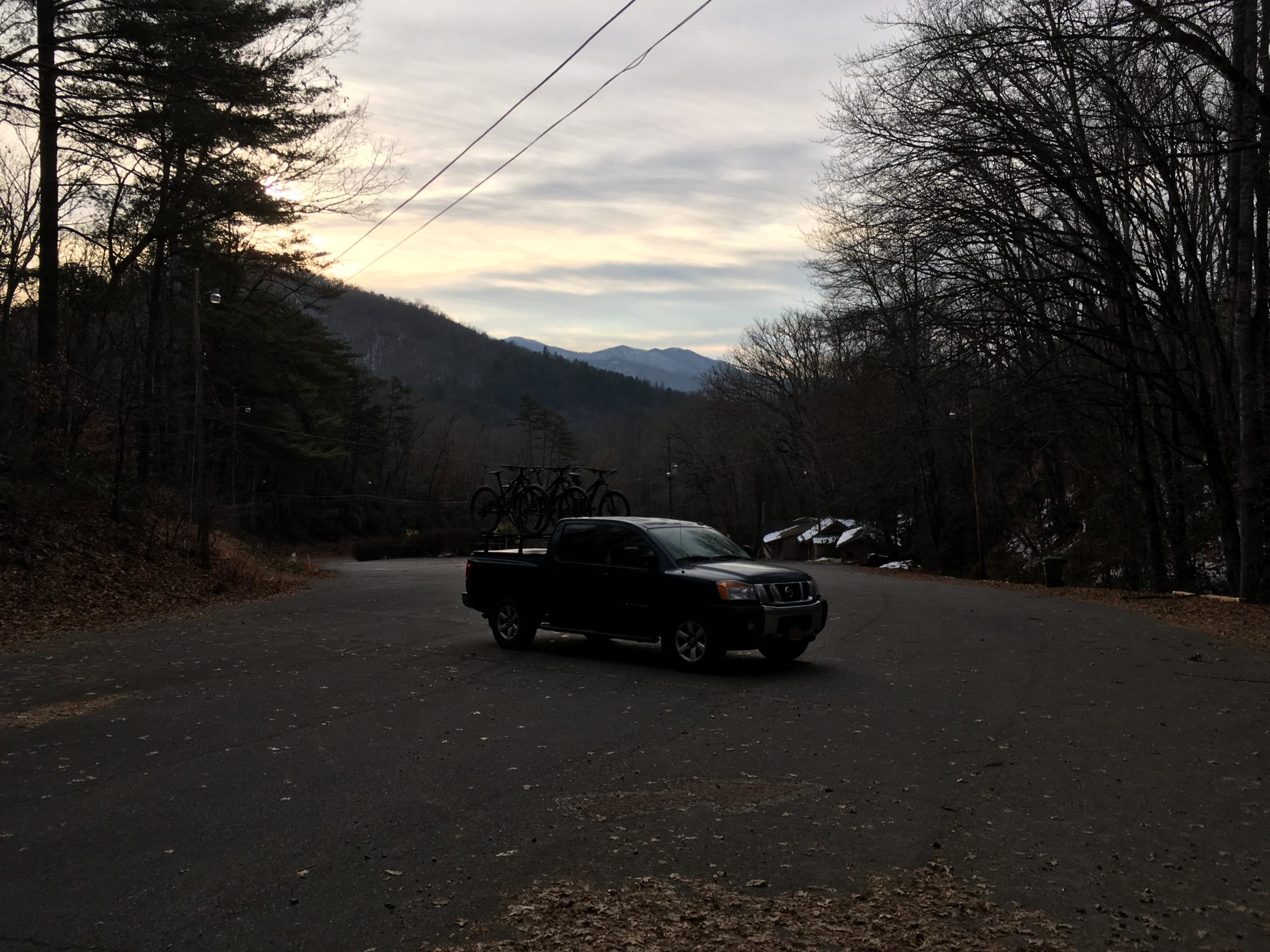 A black pickup truck parked on a winding road surrounded by trees, with a backdrop of distant mountains under a cloudy sky. The truck has bicycles mounted on the roof rack, and scattered autumn leaves cover the ground. Fire Mountain Trail System mountain bike trail.