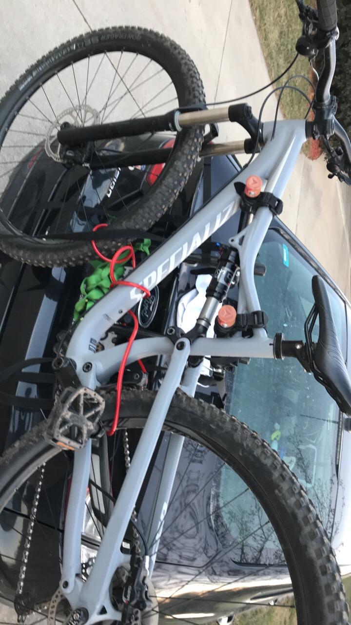 Specialized Camber: A gray mountain bike is resting on the roof of a black car. The bike is secured with straps, featuring knobby tires and a lightweight frame with visible suspension components. The scene is set outdoors, with grass in the background and a clear view of the vehicle's windshield.