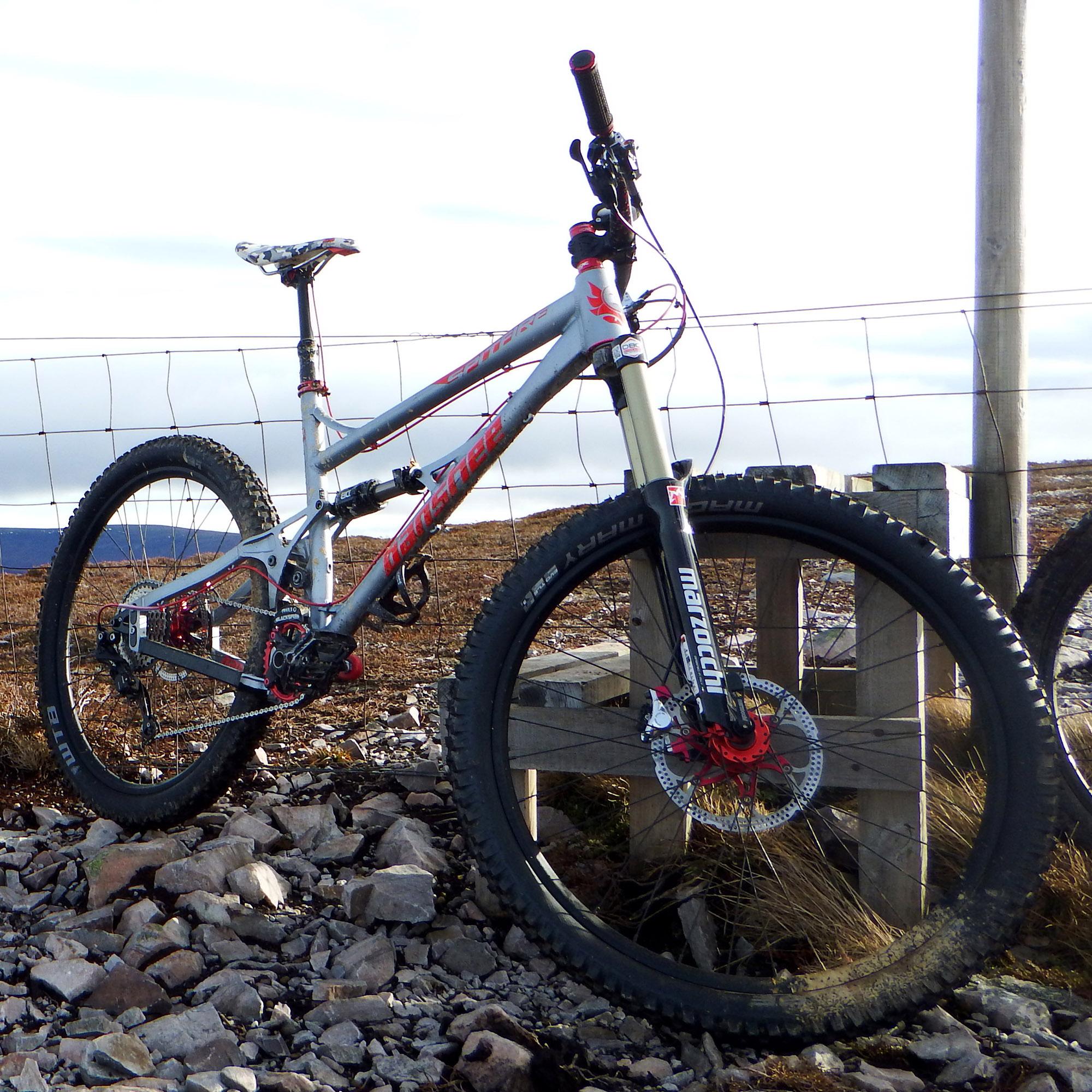 Banshee Spitfire: A mountain bike resting on rocky terrain, featuring a gray and red frame, wide tires, and visible disc brakes. The bike is positioned next to a wooden structure with a fence in the background. The landscape is open and hilly under a partly cloudy sky.