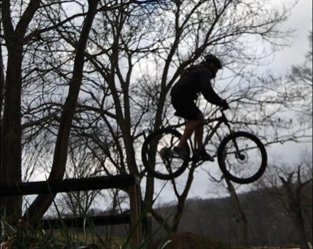 A person wearing a helmet and cycling gear jumps off a wooden ramp on a mountain bike, surrounded by bare trees under a cloudy sky. Two Rivers Bike Park mountain bike trail.