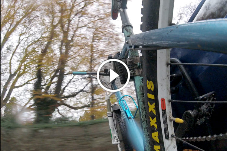 A close-up view of a mountain bike's frame, tire, and handlebars, with trees in the background blurred to convey motion. The image emphasizes the bike's components, including a prominently displayed "Maxxis" tire. A play button overlay suggests the image is linked to a video.