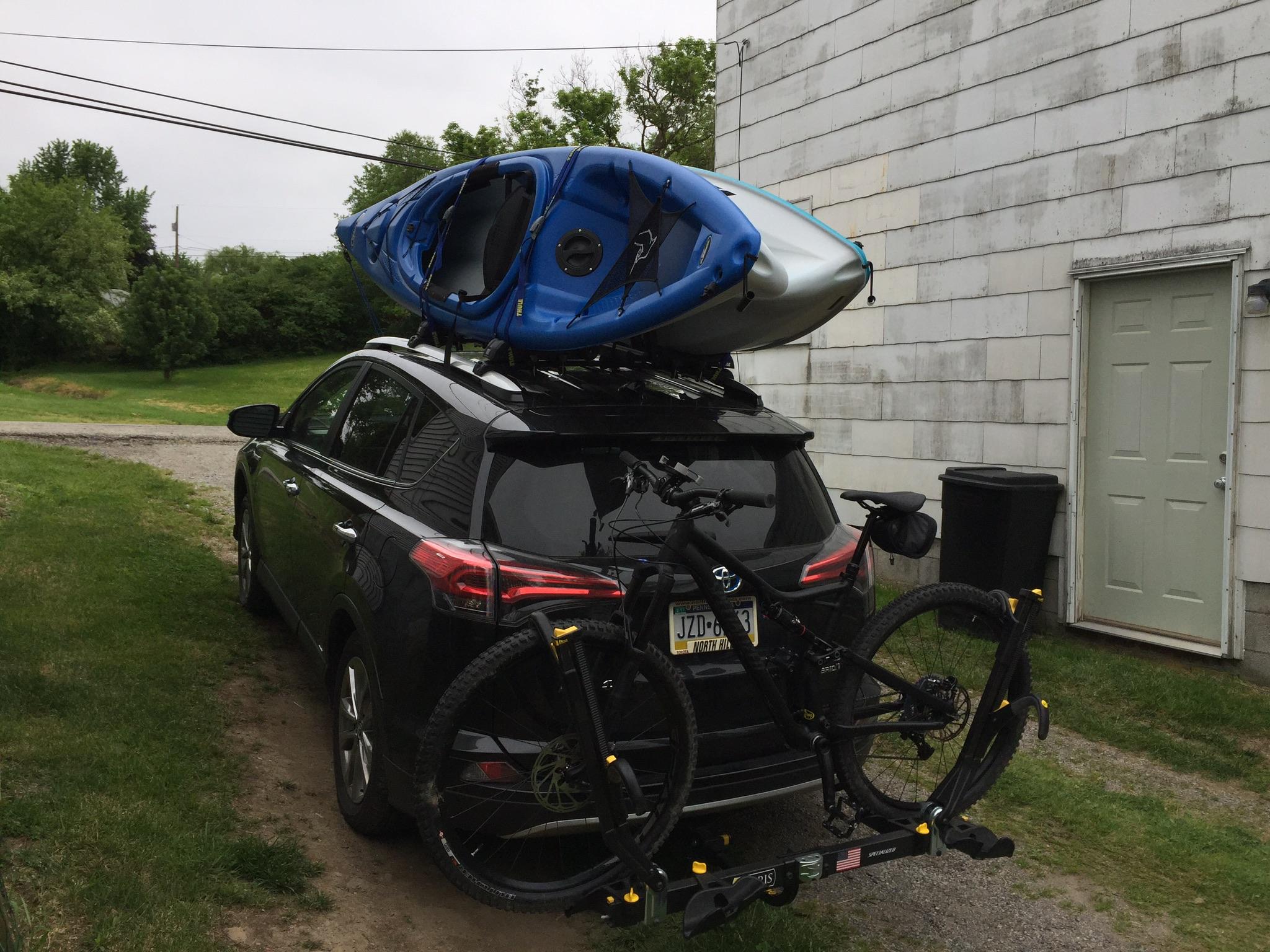 Specialized Stumpjumper FSR: A black SUV parked on a grassy area with a blue kayak secured on the roof and a mountain bike attached to the rear rack. In the background, there is a grey, weathered wall and a door. The scene is set on a cloudy day with green trees surrounding the area.