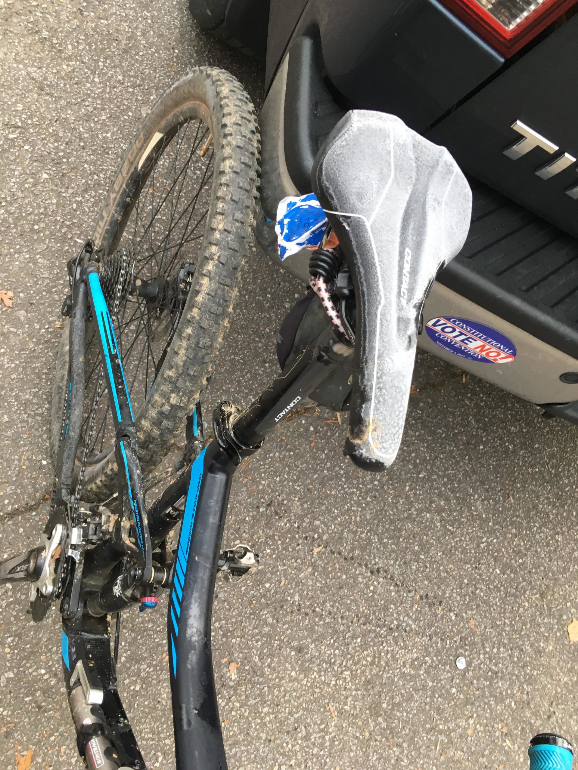 Image of a mountain bike resting against a vehicle, showing a close-up of the bike's seat and part of the frame. The bike has a slightly muddy tire, indicating off-road use. The background includes a portion of a car with a political sticker. The surface is an asphalt road with some scattered leaves. Fire Mountain Trail System mountain bike trail.
