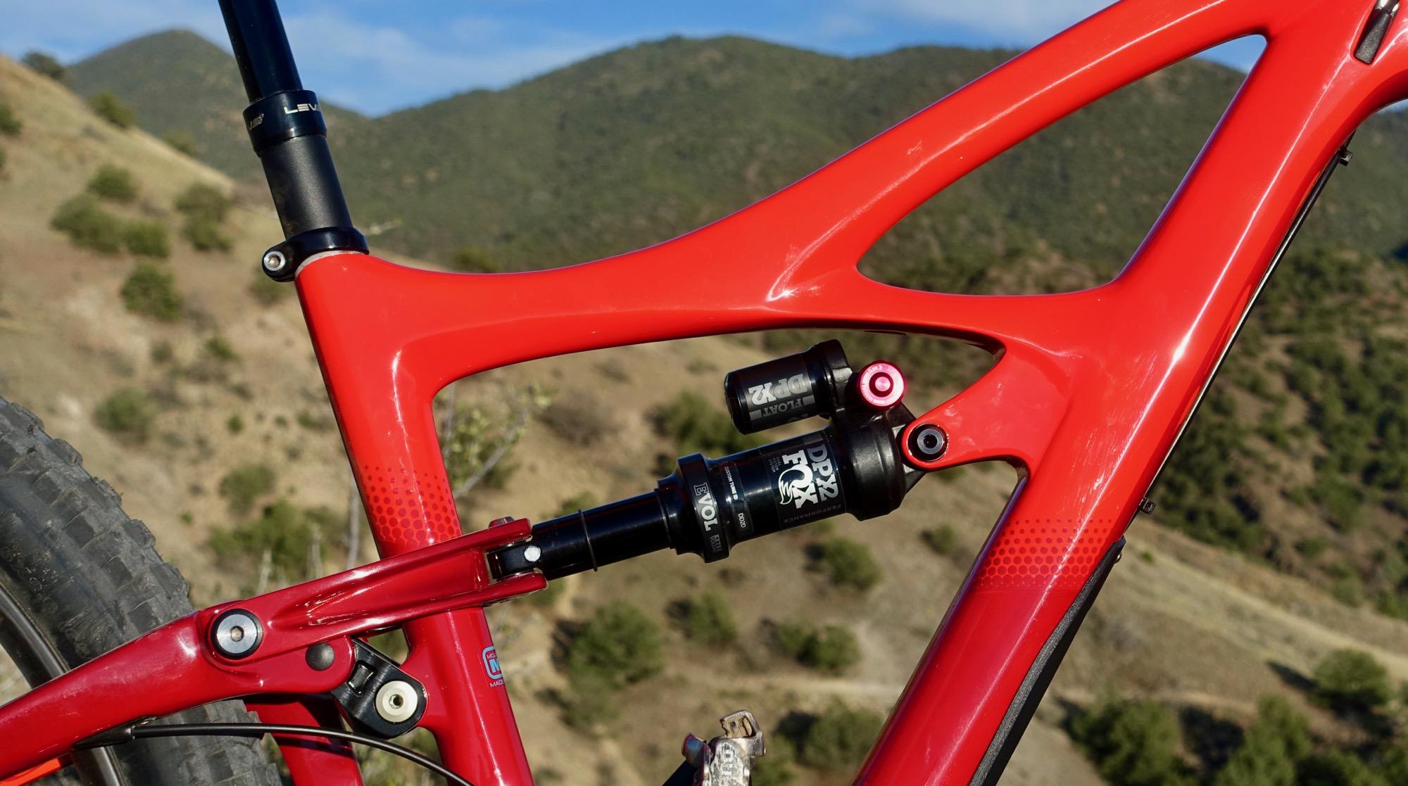 Ibis Mojo HD4: Close-up of a red bicycle frame showcasing the suspension system, including a black shock absorber, set against a mountainous background.