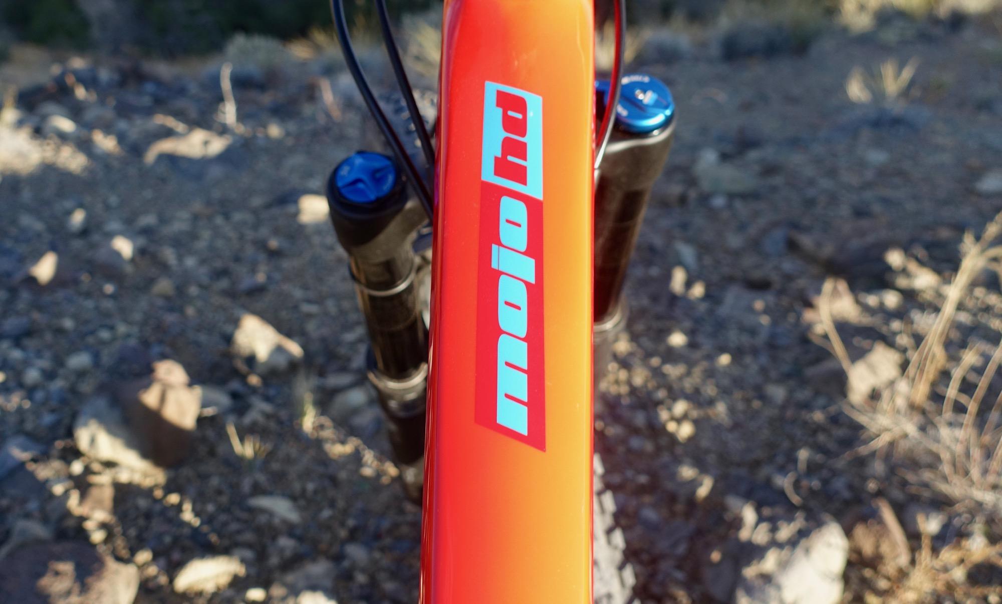 Ibis Mojo HD4: Close-up view of a mountain bike frame with the "mojo hd" logo prominently displayed. The bike is positioned on rocky terrain, showcasing its vibrant orange color and blue accents on the suspension components.
