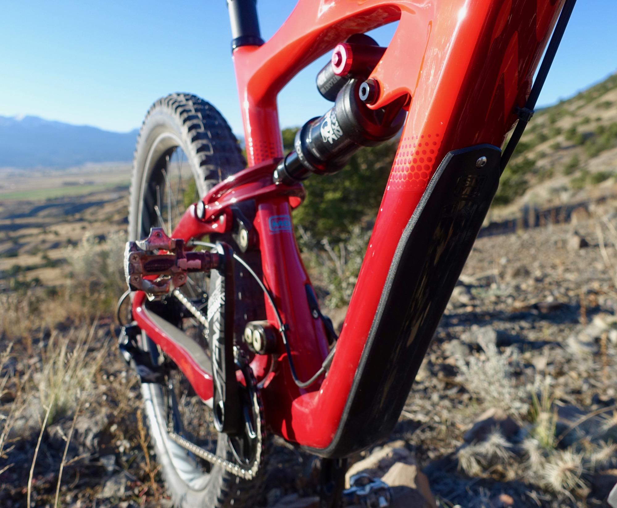 Ibis Mojo HD4: A close-up view of a red mountain bike frame with a focus on the rear section, showcasing the chain, pedals, and suspension components. The background depicts a mountainous terrain under a clear blue sky, with greenery and rocky ground visible.
