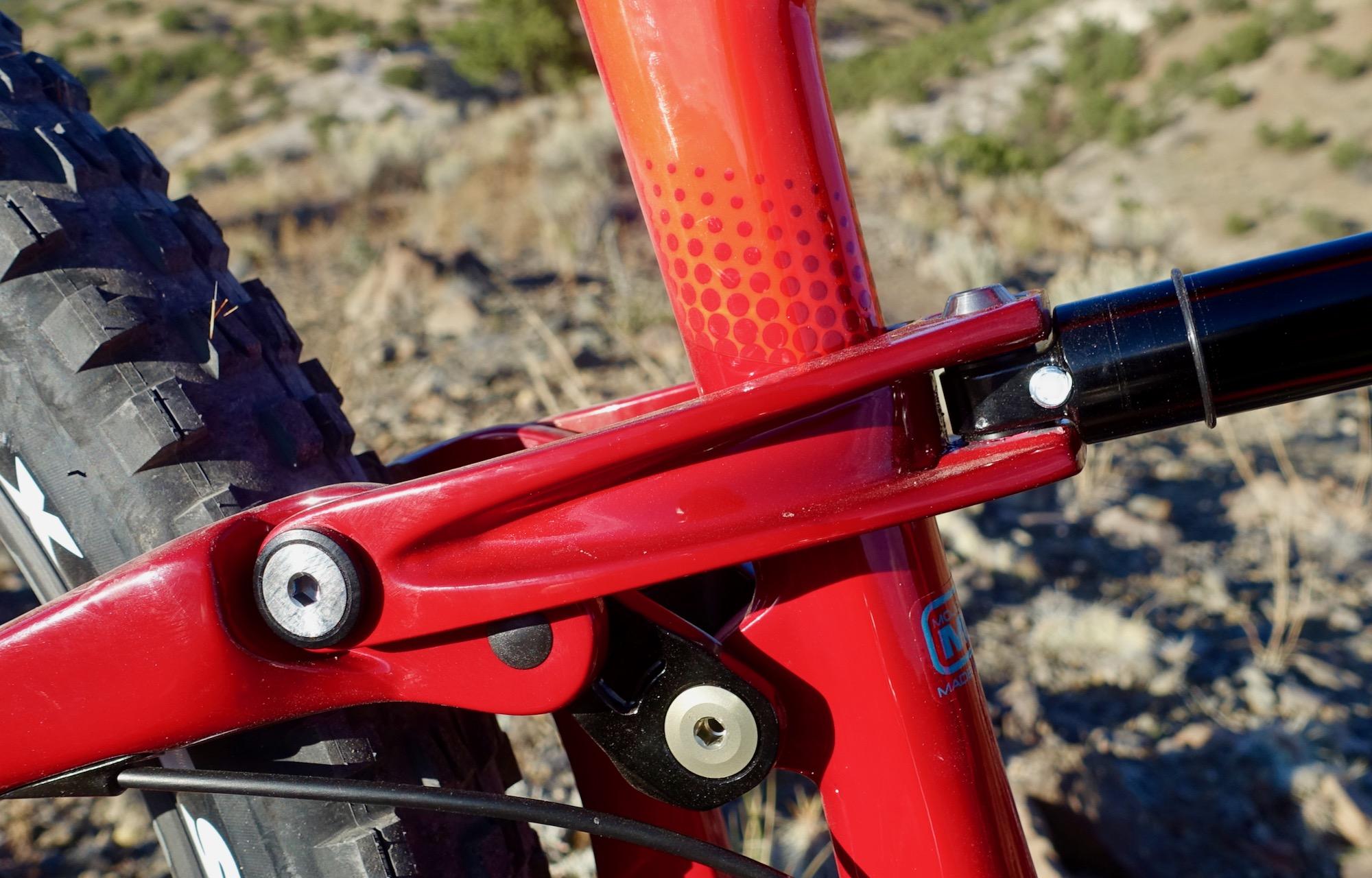 Ibis Mojo HD4: Close-up view of a red mountain bike frame featuring a detailed shot of the linkage and shock mount system. The image captures the textured surface and color gradient of the bike's frame, showcasing the mechanical components and tire treads in the background, highlighting the rugged outdoor setting.