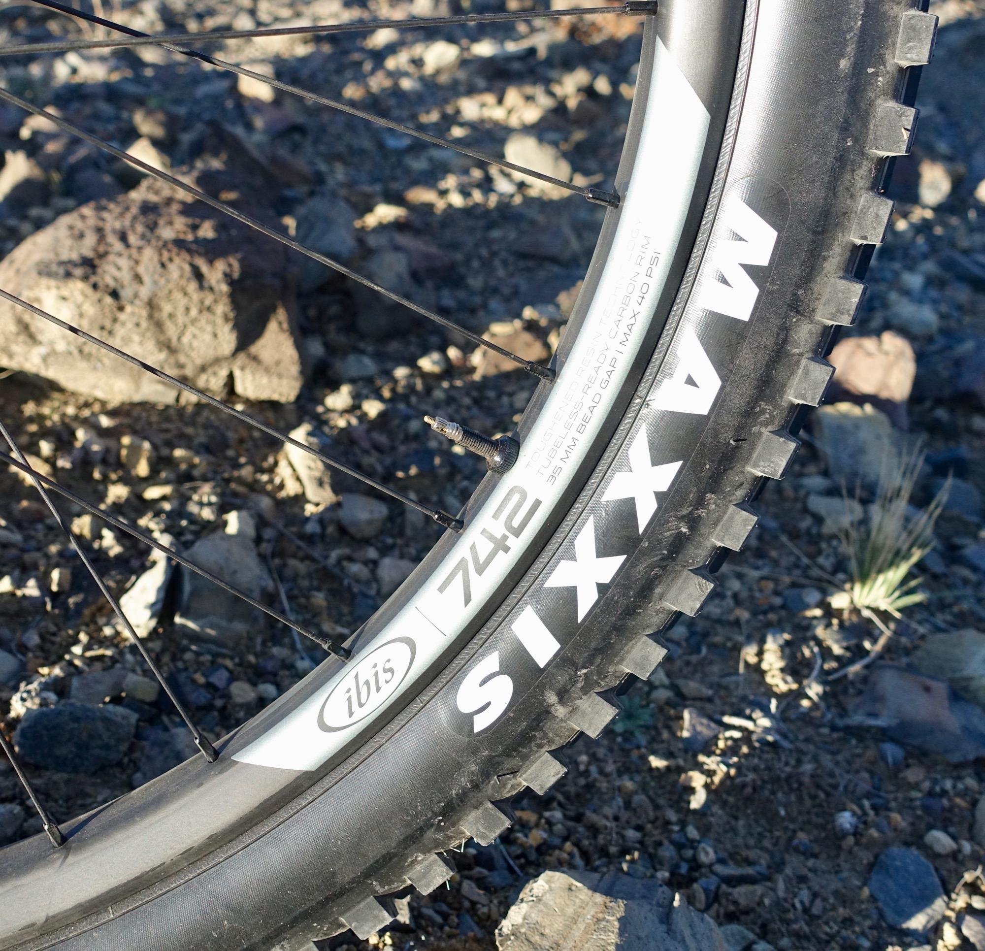 Ibis Mojo HD4: Close-up of a mountain bike tire mounted on an ibís 742 rim, featuring the Maxxis tire brand. The background consists of rocky terrain with some grassy elements, highlighting an outdoor cycling environment.