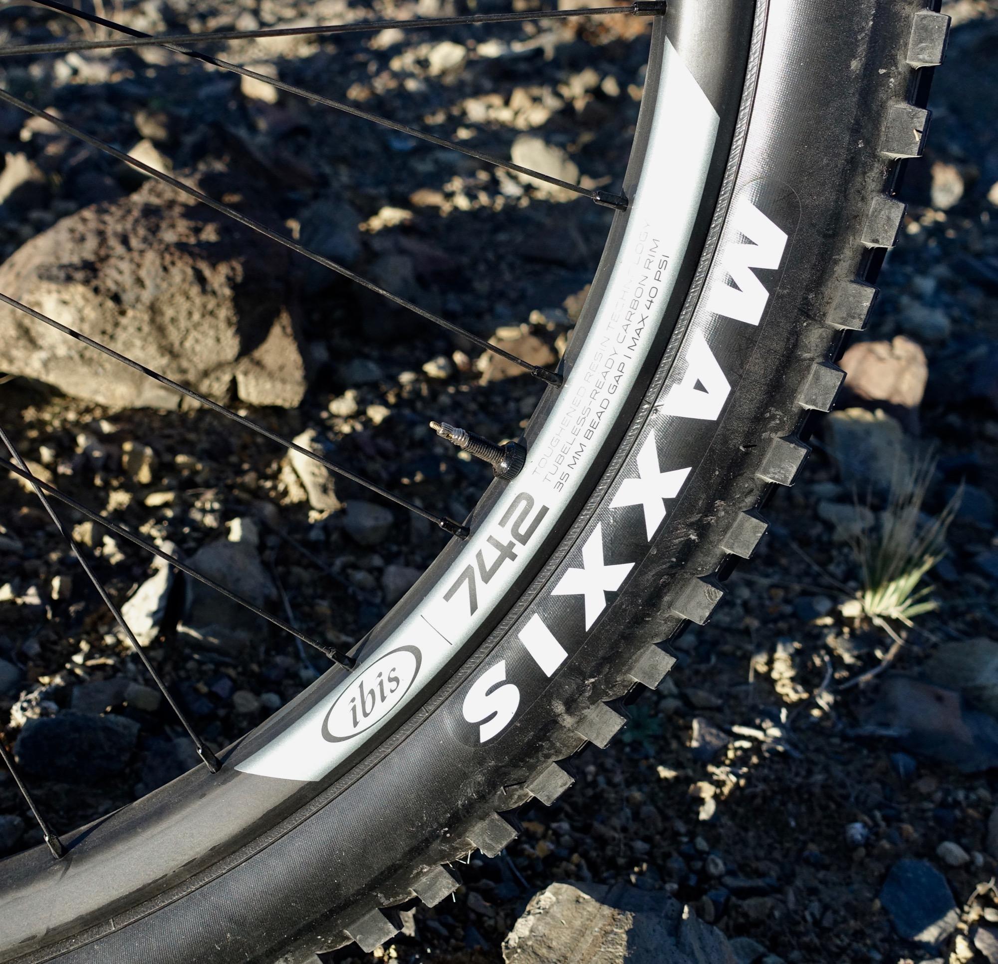 Ibis Mojo HD4: Close-up image of a mountain bike tire showing the "Ibis 742" and "Maxxis Six" branding, with visible knobby tread patterns, on a rocky terrain background. The tire's valve and spokes are also partially visible.