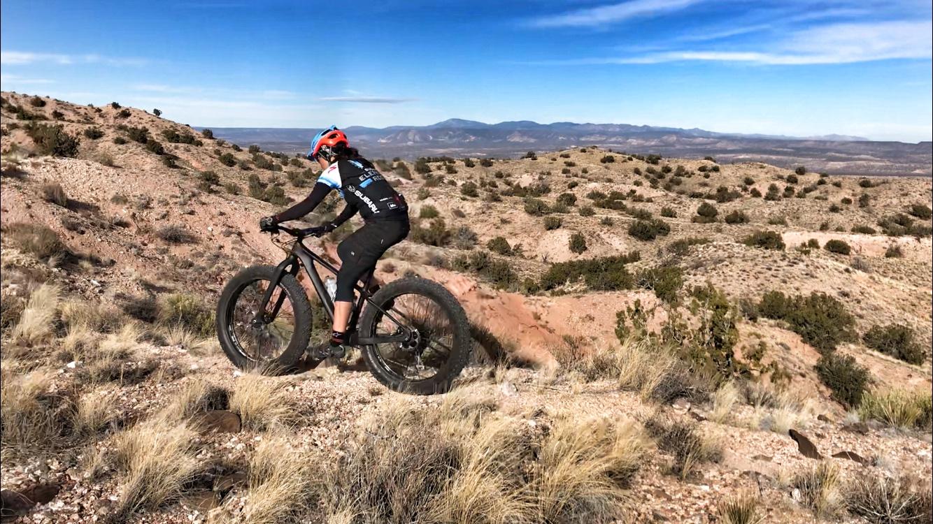 A cyclist riding a fat bike on a rocky trail in a desert landscape, surrounded by sparse vegetation and distant mountains under a clear blue sky. Mariposa Fat Bike Trails mountain bike trail.