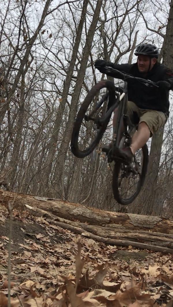 A mountain biker airborne over a log on a dirt trail, surrounded by bare trees and fallen leaves, showcasing an action shot in a forested area. Petrifying Springs mountain bike trail.