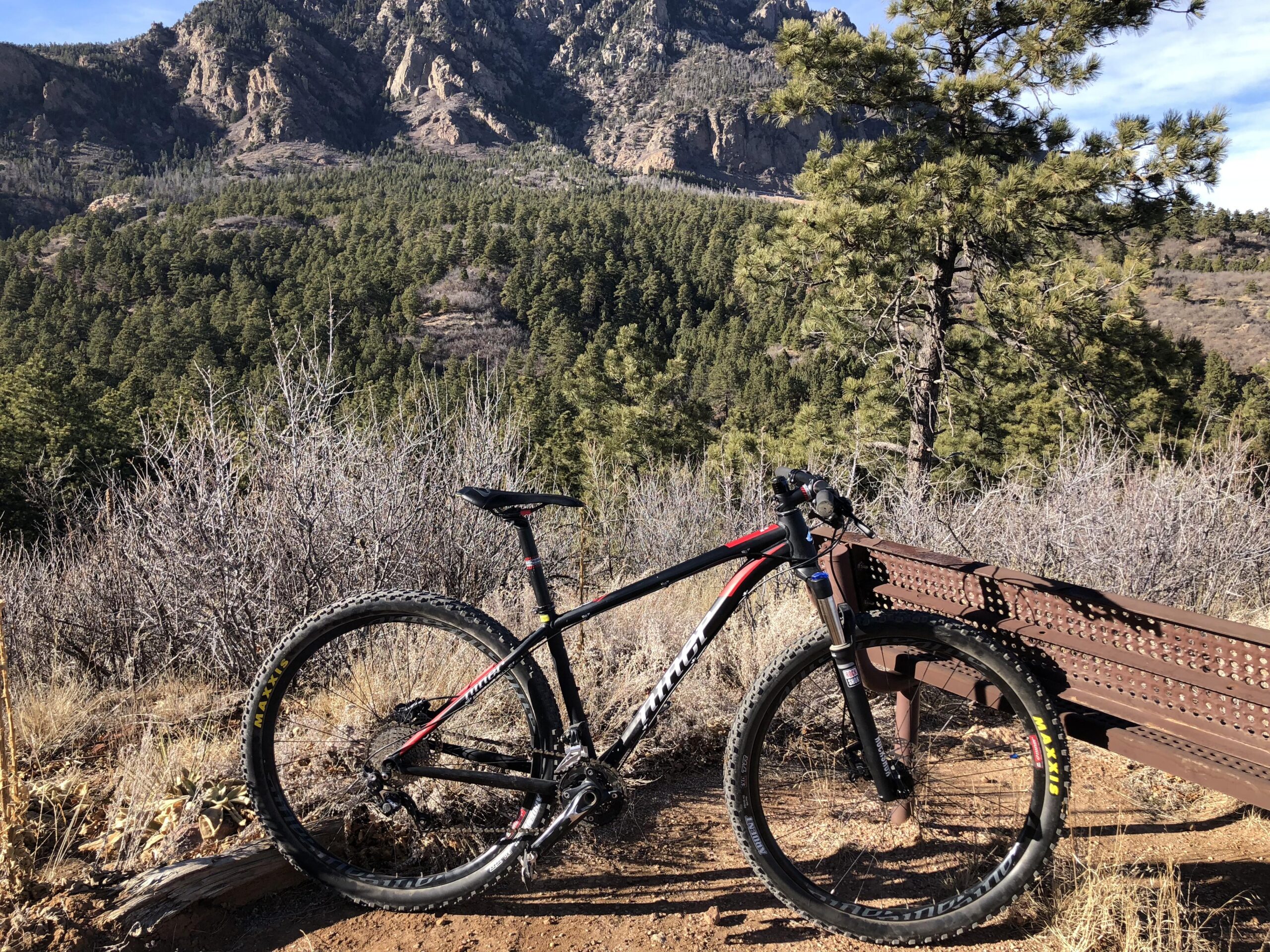 Niner EMD 9: A mountain bike rests on a wooden bench overlooking a scenic landscape of mountains and dense trees, under a clear blue sky. The bike features a black frame with red accents and is positioned on a dirt path surrounded by dry vegetation.