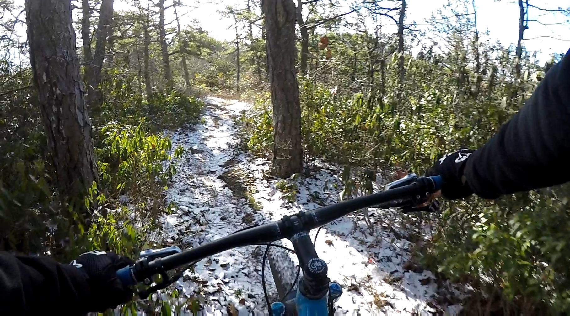A mountain biker's perspective while riding on a narrow, snow-dusted trail through a forested area, surrounded by trees and greenery. The handlebars of the bike are visible, with gloved hands gripping them as the trail winds ahead. North Mountain/longdale Loop mountain bike trail.
