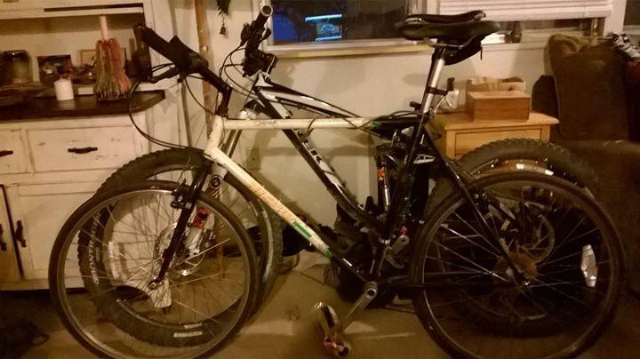 Trek Remedy 8: Two bicycles are parked closely together inside a room. The first bike is white with green accents, and the second is black. Both have thick tires suitable for off-road cycling. The background shows a wooden cabinet and a couch, with dim lighting creating a cozy atmosphere.