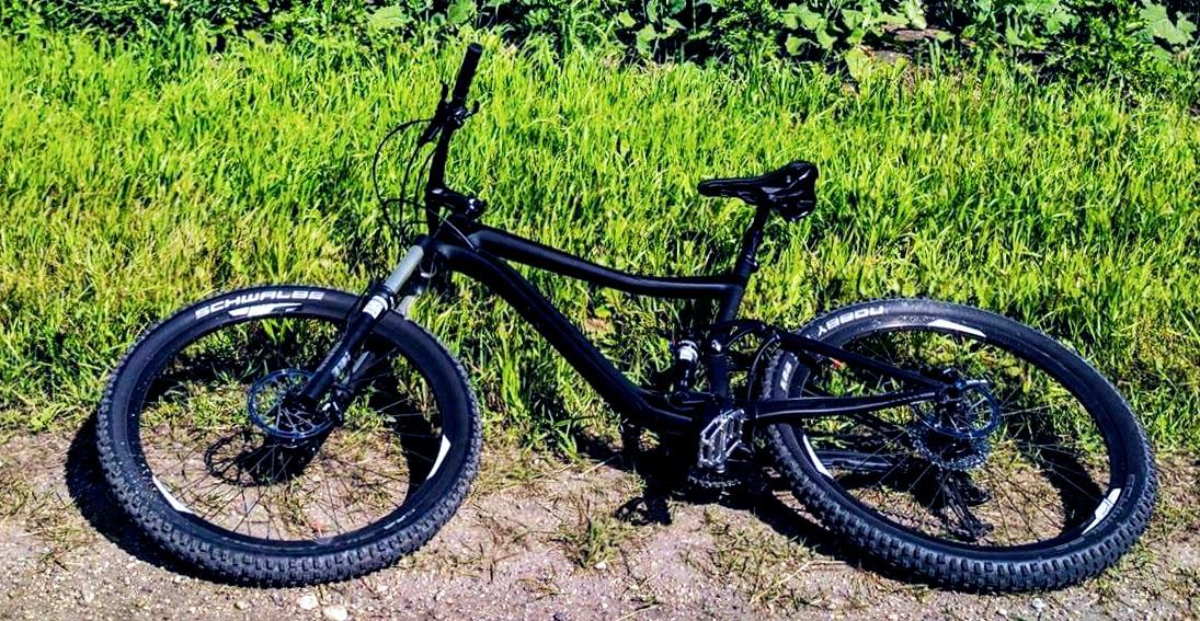 Giant Giant Trance Advanced 27.5 2: A black mountain bike lying on a gravel path beside a lush green field. The bike features thick tires suitable for rough terrains and has a curved frame for stability and comfort.