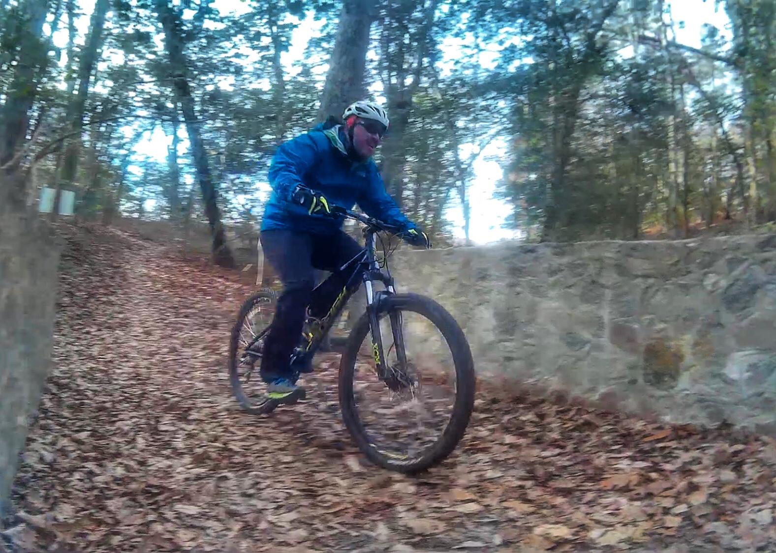 A mountain biker in a blue jacket rides along a leaf-covered trail in a wooded area. Trees surround the path, and a stone wall is partially visible to the right. The cyclist appears focused and is wearing a helmet for safety. Appomattox River Regional Park Trail mountain bike trail.