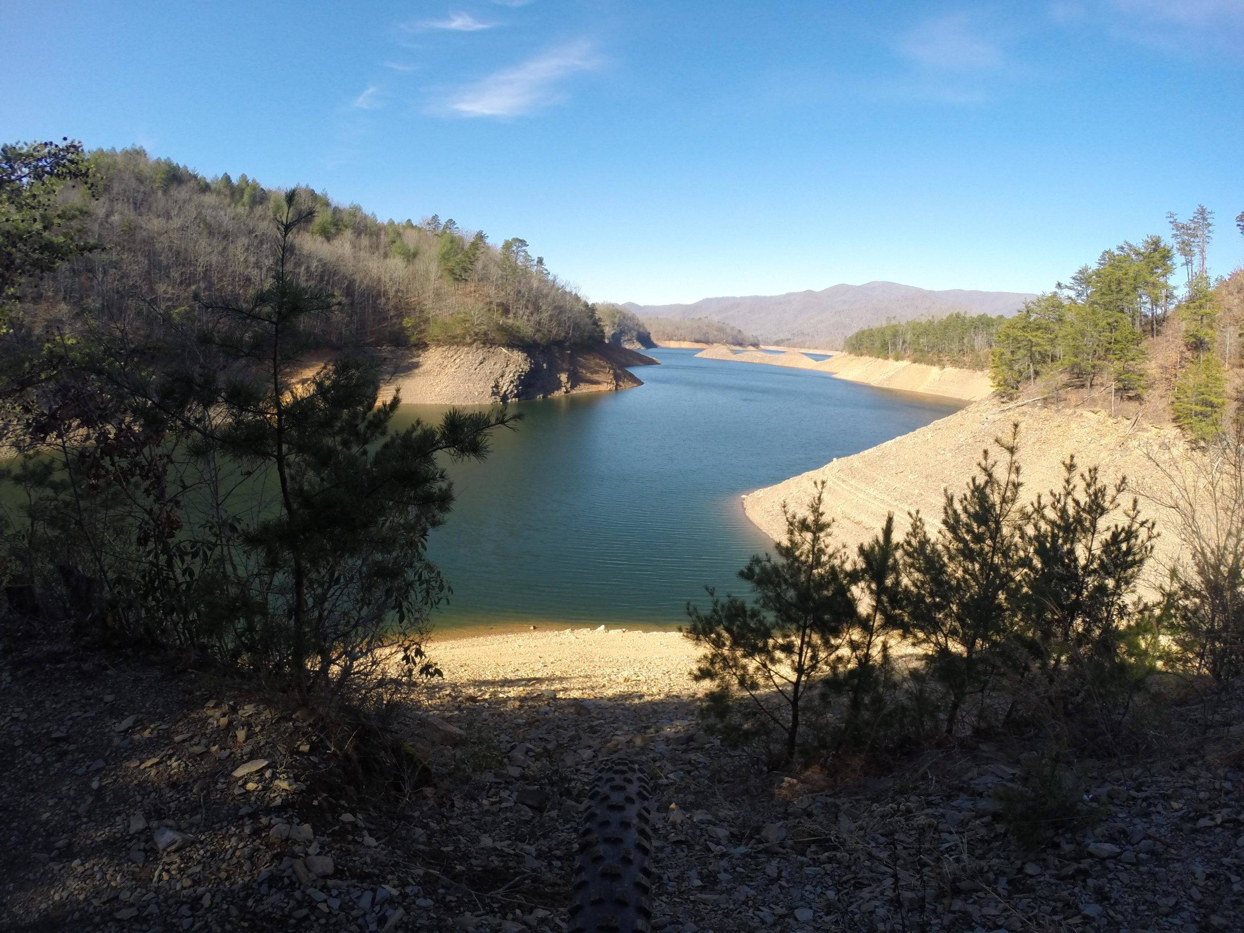 A serene landscape featuring a calm lake surrounded by gently sloping hills and trees, under a clear blue sky. The shoreline is visible with a mix of rocky and sandy areas, suggesting low water levels. The scene conveys a tranquil natural setting. Tsali Recreation Area mountain bike trail.