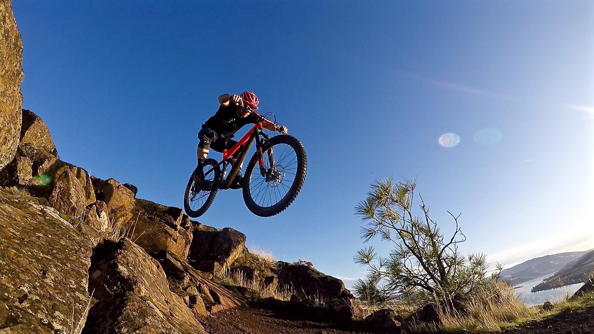 A mountain biker in mid-air performing a jump over rocky terrain, with a clear blue sky in the background and a landscape featuring a body of water and hills. Syncline mountain bike trail.