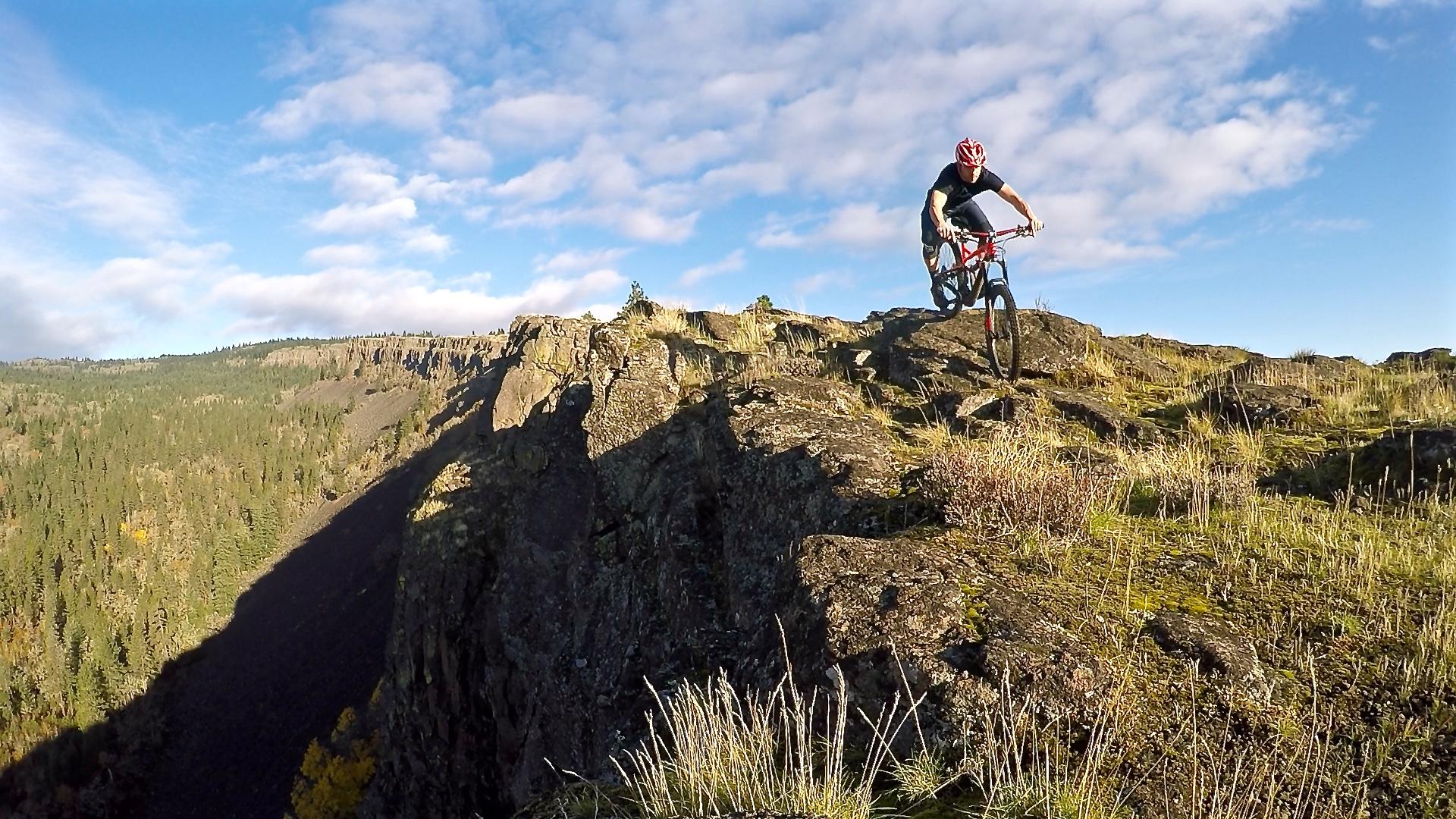 A mountain biker rides along the edge of a rocky cliff surrounded by lush green trees under a bright blue sky with fluffy clouds. The biker is in a dynamic pose, showcasing skill and agility on rugged terrain. Syncline mountain bike trail.