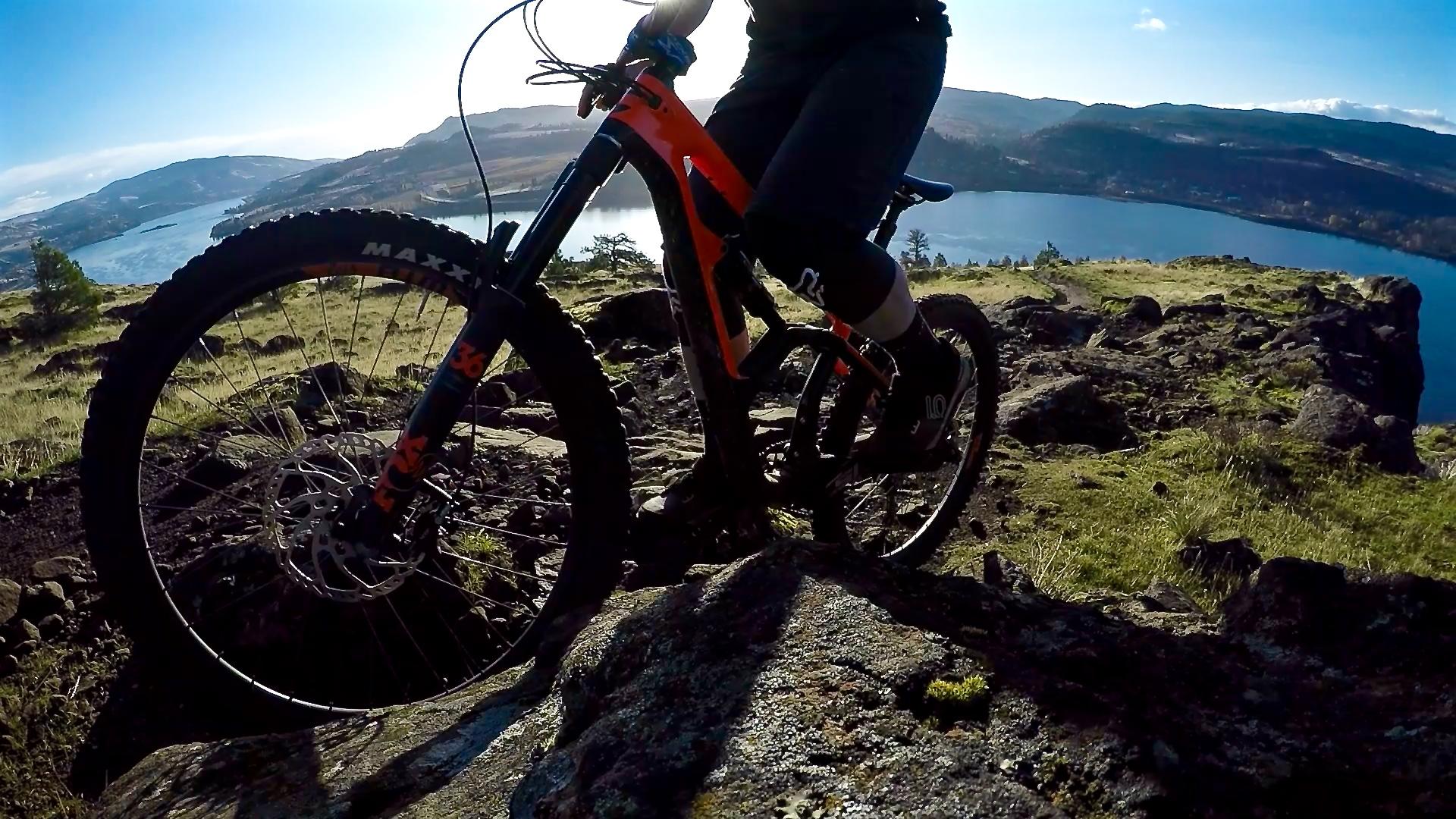 A person riding a mountain bike on rocky terrain, with one foot on a rock and a picturesque landscape featuring a lake and hills in the background under a clear blue sky. Syncline mountain bike trail.