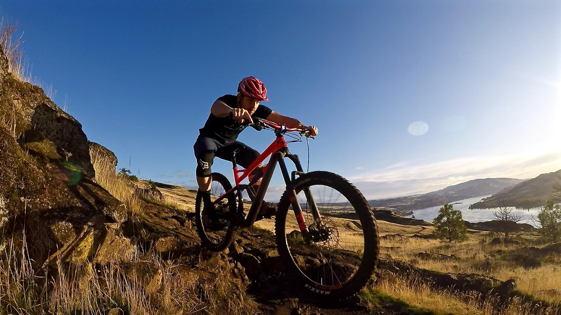 A person riding a mountain bike over rocky terrain, with a scenic landscape featuring hills and a river in the background. The cyclist is wearing a helmet and athletic gear, showcasing an outdoor adventure in bright sunlight. Syncline mountain bike trail.