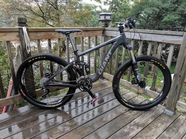 Giant Trance X1: A grey mountain bike is parked on a wooden deck, surrounded by greenery. The bike features distinctive branding along the frame and has thick tires suitable for off-road riding. Rain droplets are visible on the deck, indicating recent wet weather.