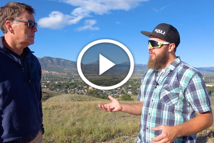 Two men are standing on a hillside with a mountainous landscape in the background. One man is wearing sunglasses and a blue plaid shirt, speaking animatedly while using hand gestures. The other man, who is wearing sunglasses and a dark sweater, appears to be listening intently. A play button is overlaid on the image, indicating it is a video.