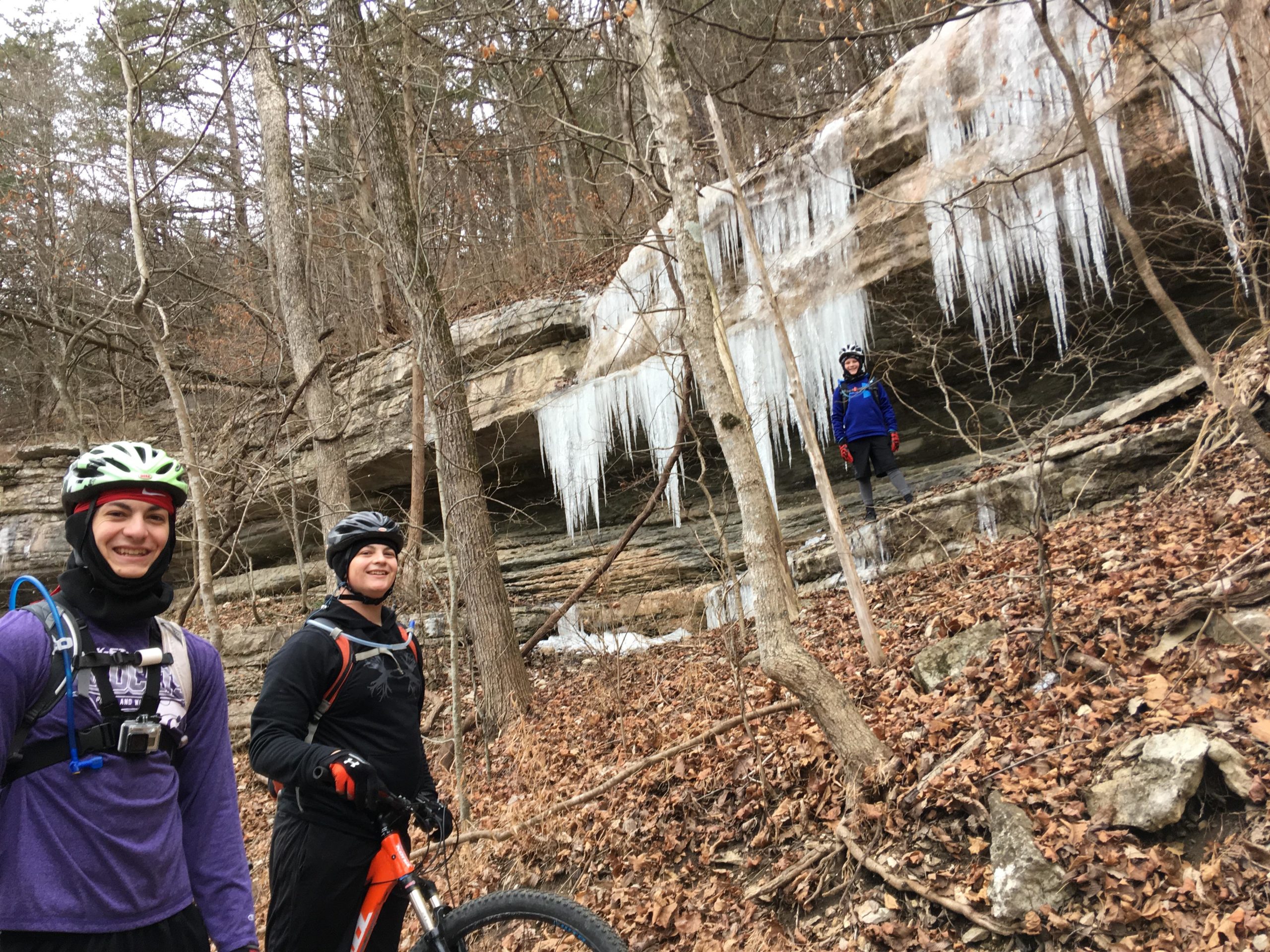 Three mountain bikers pose for a photo in a forested area with icicles hanging from a rocky outcrop in the background. The two friends in the foreground are smiling, dressed in winter cycling gear and helmets, while a third person stands on the rocks behind them. The ground is covered with fallen leaves, and the trees are bare, indicating a chilly season. Blowing Springs mountain bike trail.