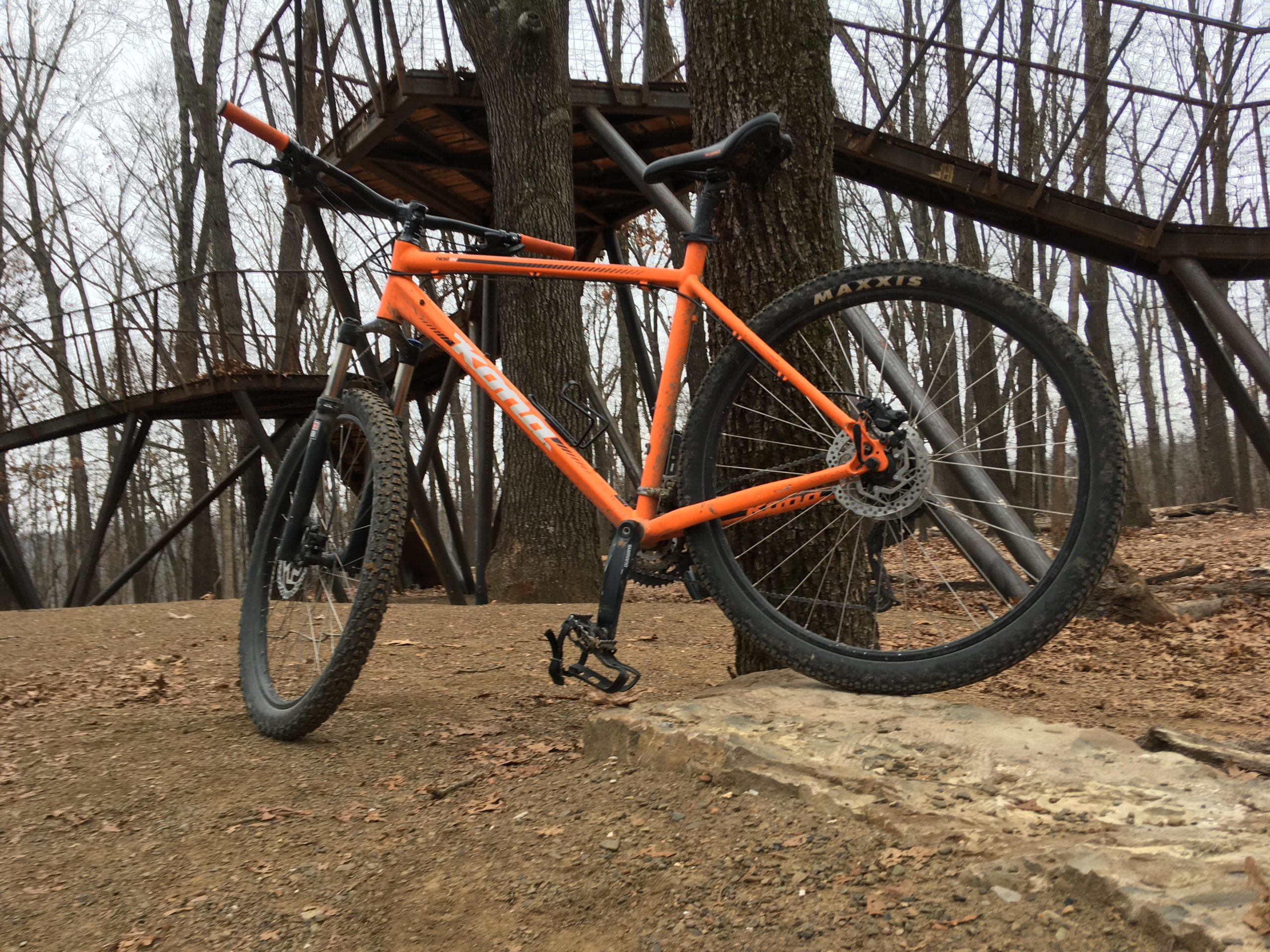 Mountain bike propped against a tree, with a wooden structure in the background. The bike features an orange frame, rugged tires, and is positioned on a dirt path surrounded by bare trees. Coler Mountain Bike Preserve mountain bike trail.