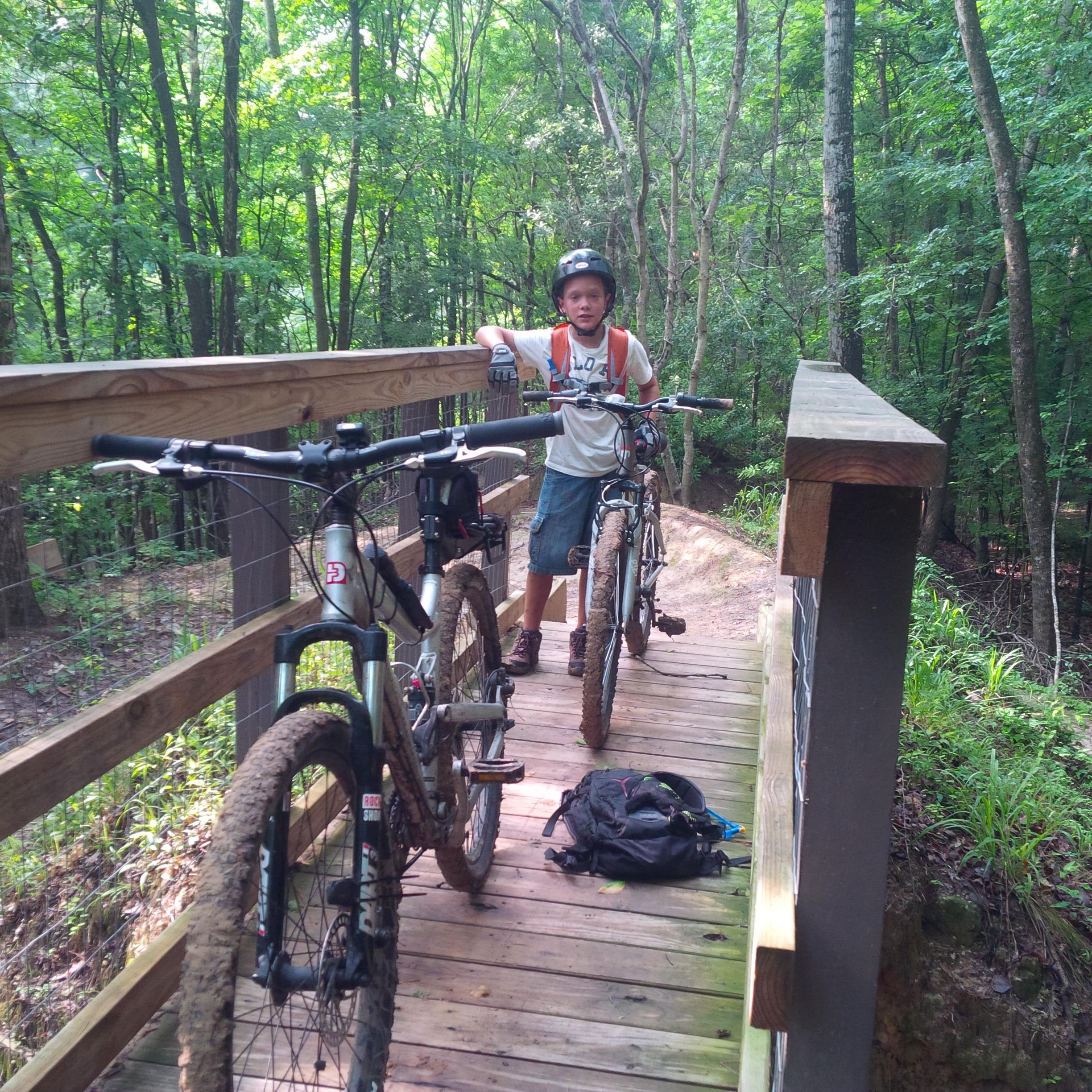 A young boy in a helmet and gloves stands on a wooden bridge in a wooded area, leaning on his mountain bike. Another bike is propped up next to him, both showing signs of mud. A black backpack is placed on the wooden floor of the bridge, surrounded by vibrant green foliage and trees in the background. Wannamaker Park mountain bike trail.