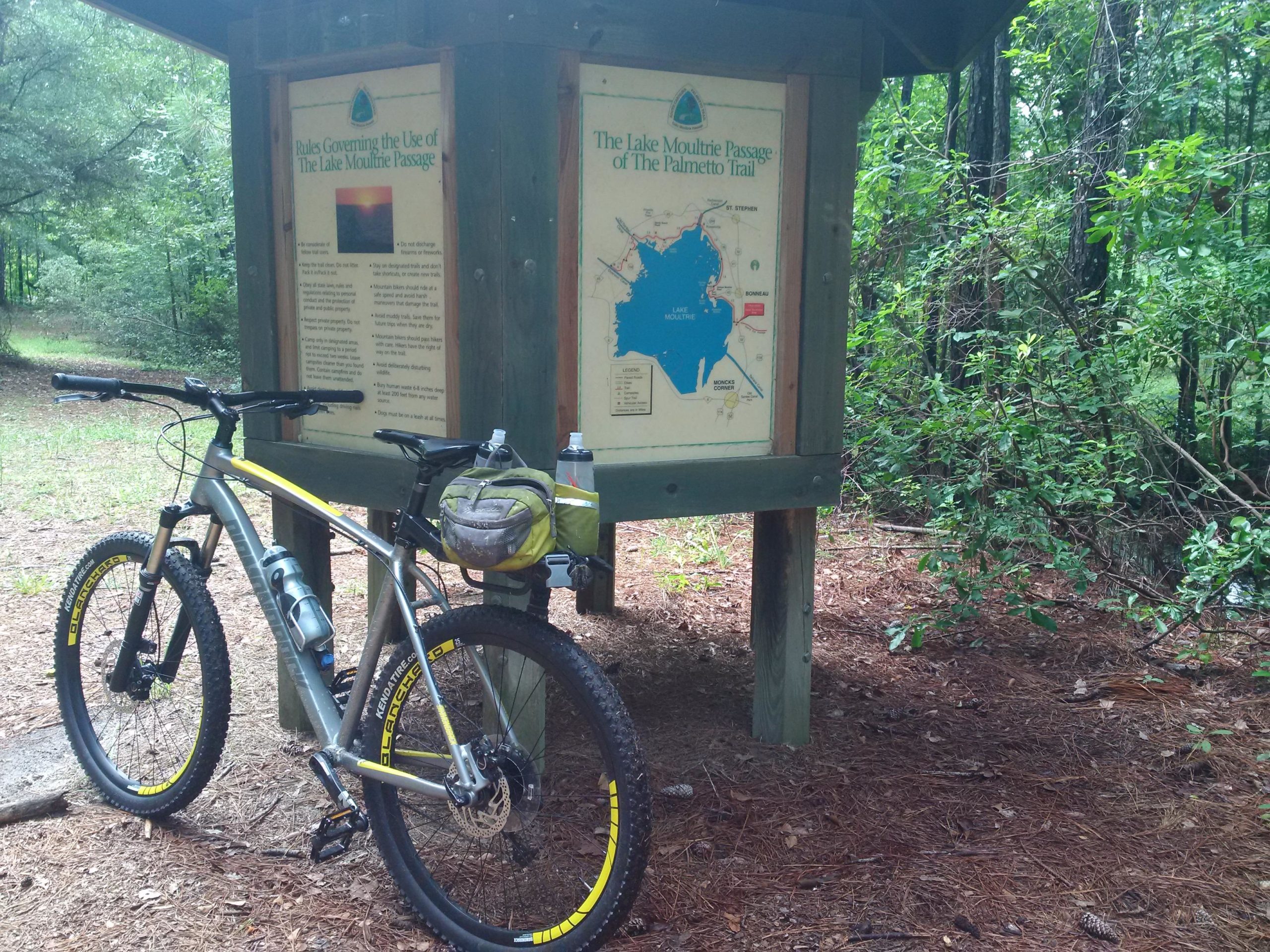 A mountain bike parked next to an informational sign about the Lake Moultrie Passage of the Palmetto Trail, surrounded by lush greenery and pine needles. The sign features rules and a map of the area. Swamp Fox Passage mountain bike trail.