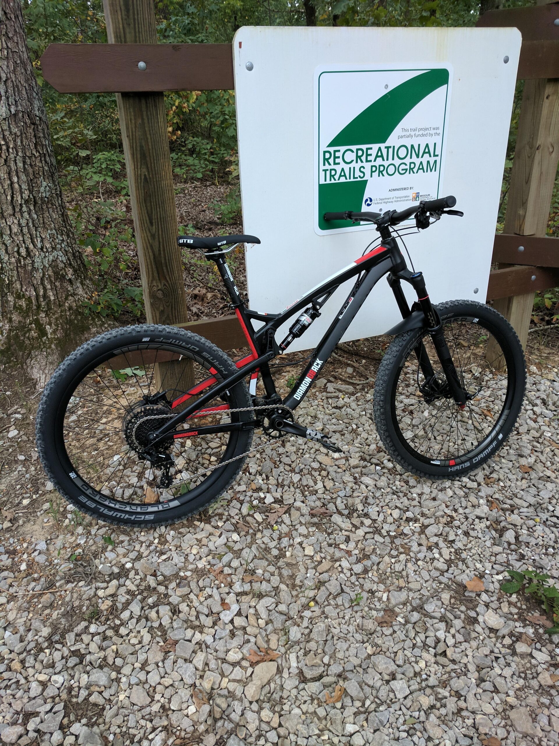 Diamondback Diamondback Release 1 27.5 2017: A black and red mountain bike parked beside a wooden sign that reads "Recreational Trails Program." The bike is positioned on a gravel path surrounded by trees with autumn foliage.