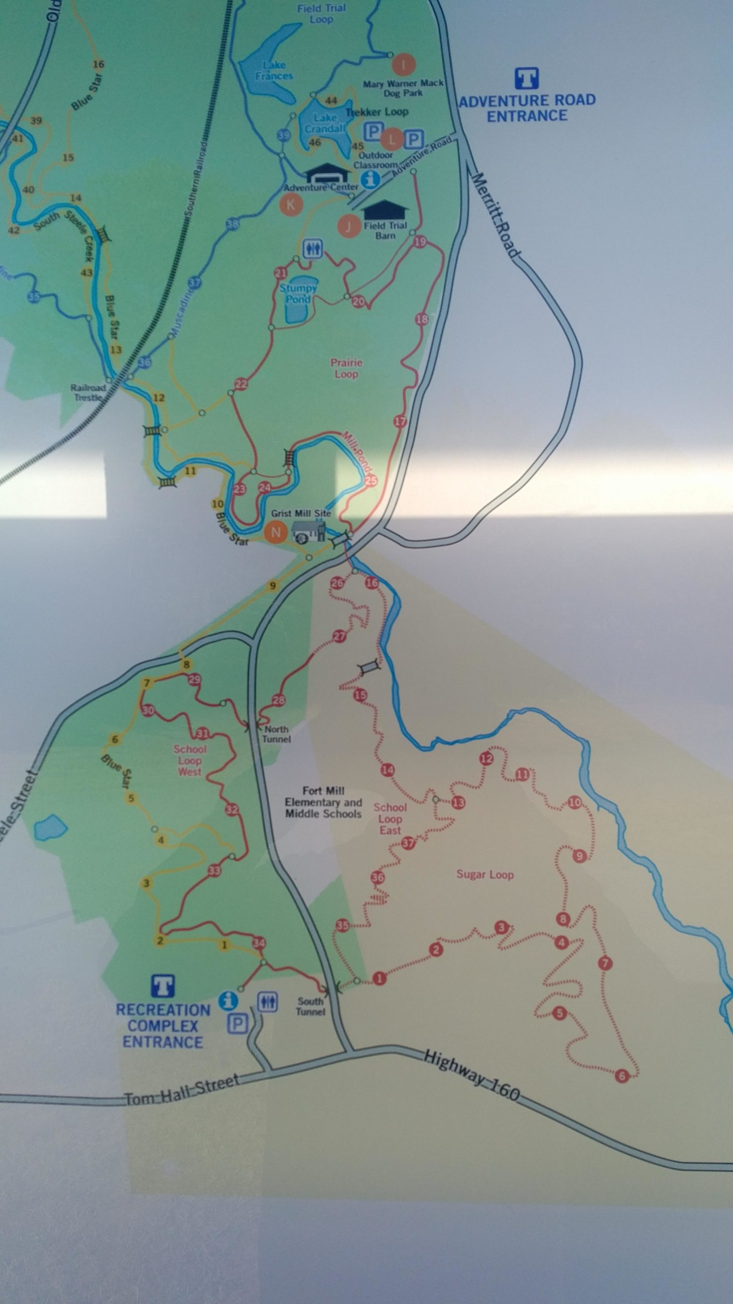 Map of a park or recreational area featuring various trails, loops, and points of interest including lakes, a dog park, and entrances to the Adventure Center and Recreation Complex. The map highlights paths for walking, biking, and other outdoor activities. Notable features include the Fort Mill schools and designated parking areas. Anne Springs Close Greenway mountain bike trail.
