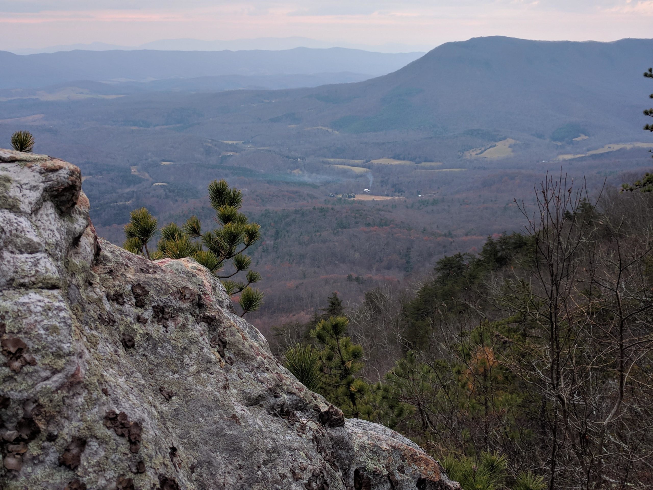 A scenic view from a rocky outcrop overlooking a mountainous landscape, with rolling hills and valleys in the distance. The foreground features a detailed rock surface and scattered pine branches, while the background showcases layered blue mountains under a cloudy sky. North Mountain/longdale Loop mountain bike trail.