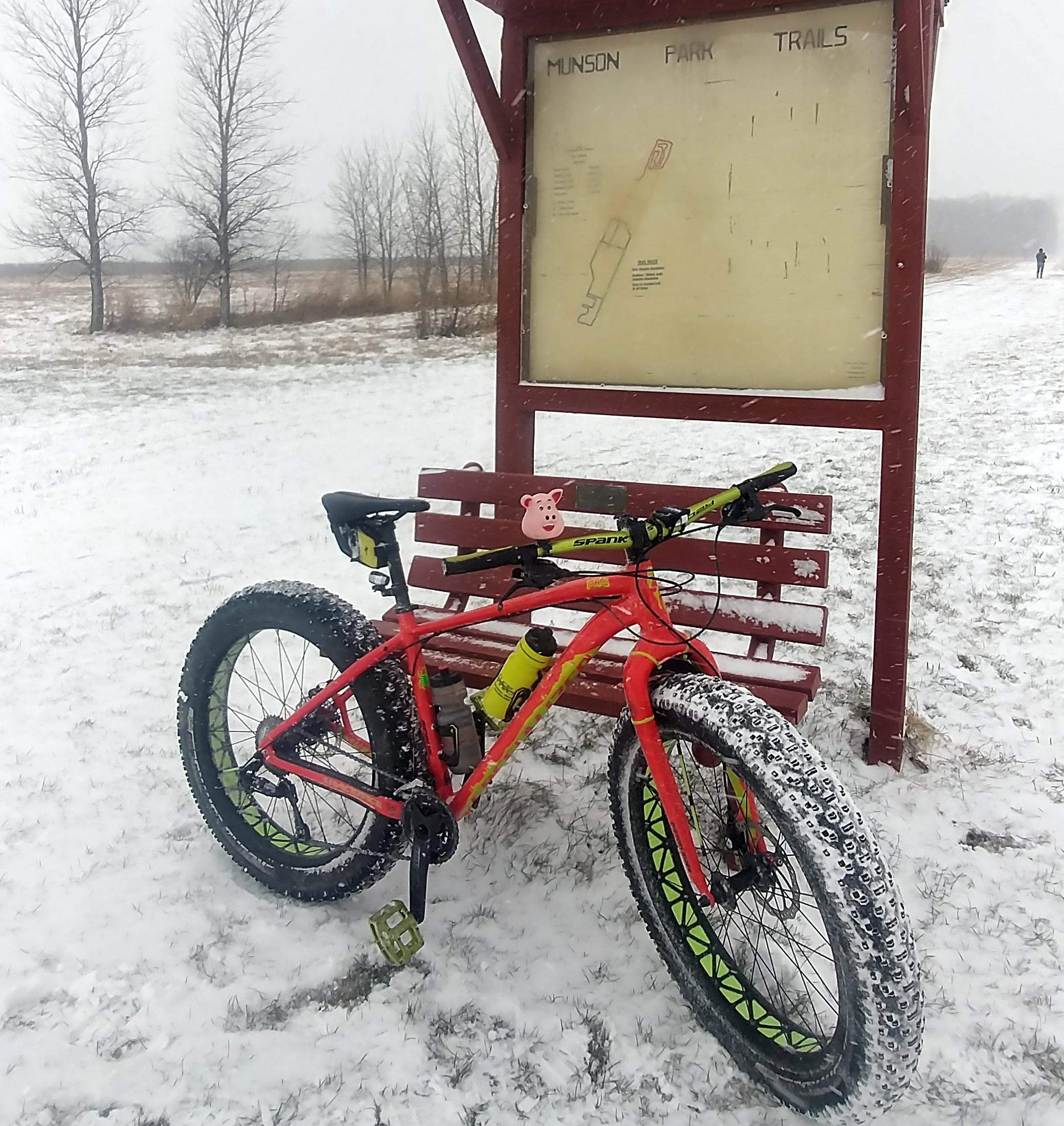 A bright red fat bike with green accents is parked on a snowy field next to a trail map sign for Munson Park. The bike features wide, heavily treaded tires, a water bottle holder, and a small pig decoration on the handlebars. In the background, bare trees are visible, and light snow is falling. Munson Park mountain bike trail.