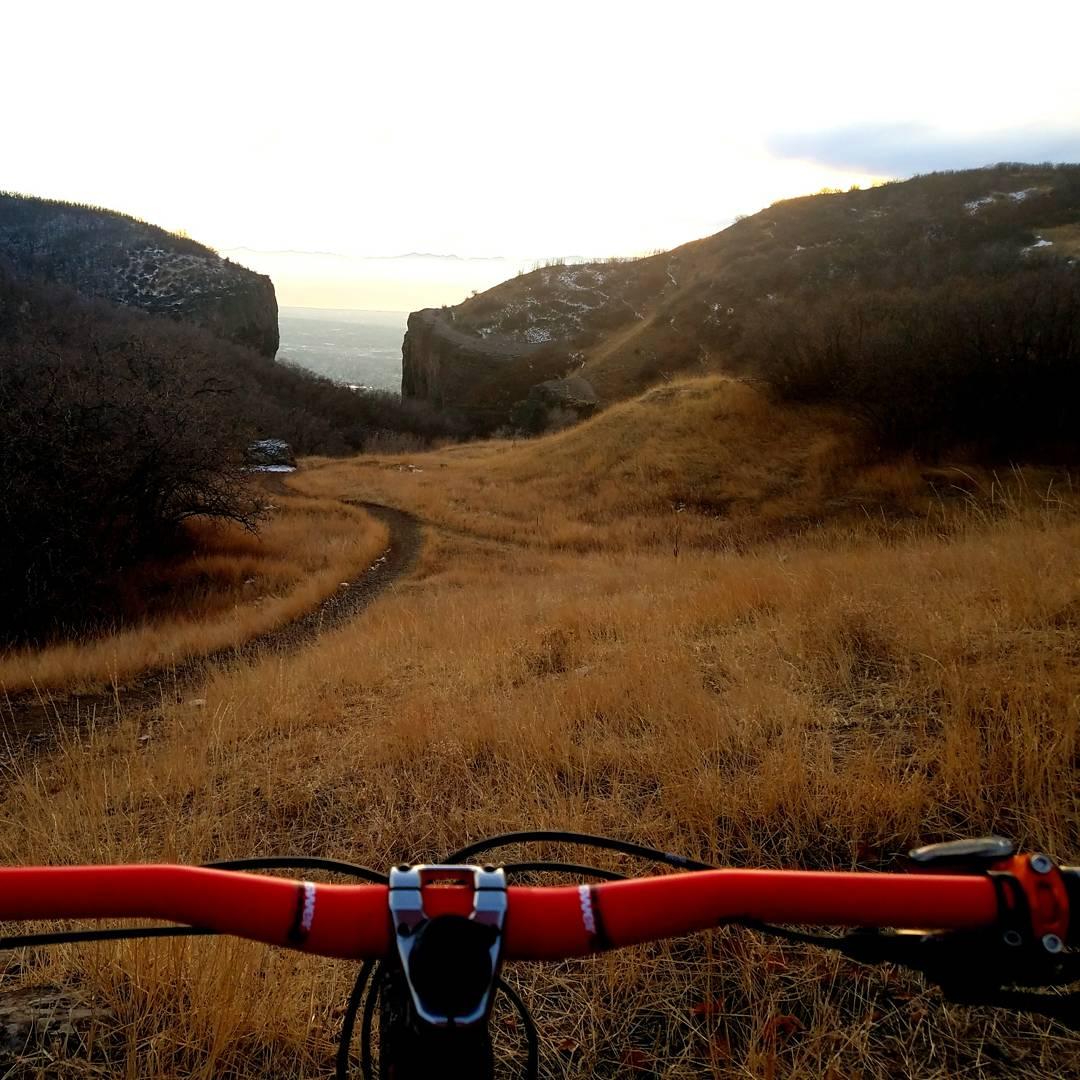A view from a mountain bike handlebars overlooking a winding dirt path through tall golden grass, with rocky cliffs and distant mountains illuminated by the soft glow of a sunset in the background. Up Shooter, Mr. Hyde, Mrs. Hyde. Down Lament, Dry Canyon DH, and Bonneville Shoreline Trail mountain bike trail.