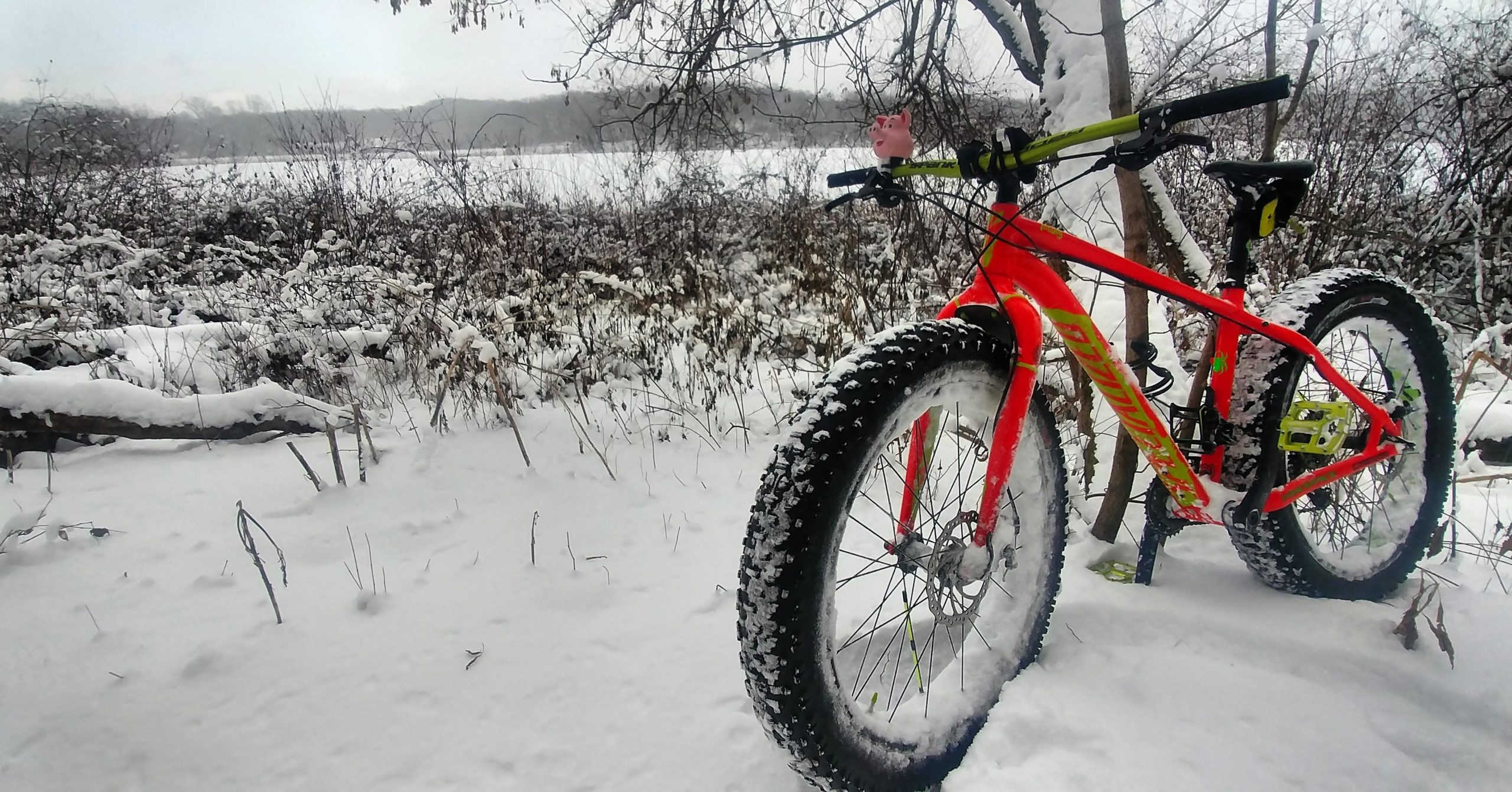 A brightly colored fat bike with oversized tires is partially buried in snow beside a snowy landscape. The bike's frame is red with green accents, and it has a small pink pig figure attached to the handlebars. Surrounding the bike is a peaceful winter scene featuring snow-covered bushes and trees in the background. DTE Energy Foundation Trail mountain bike trail.