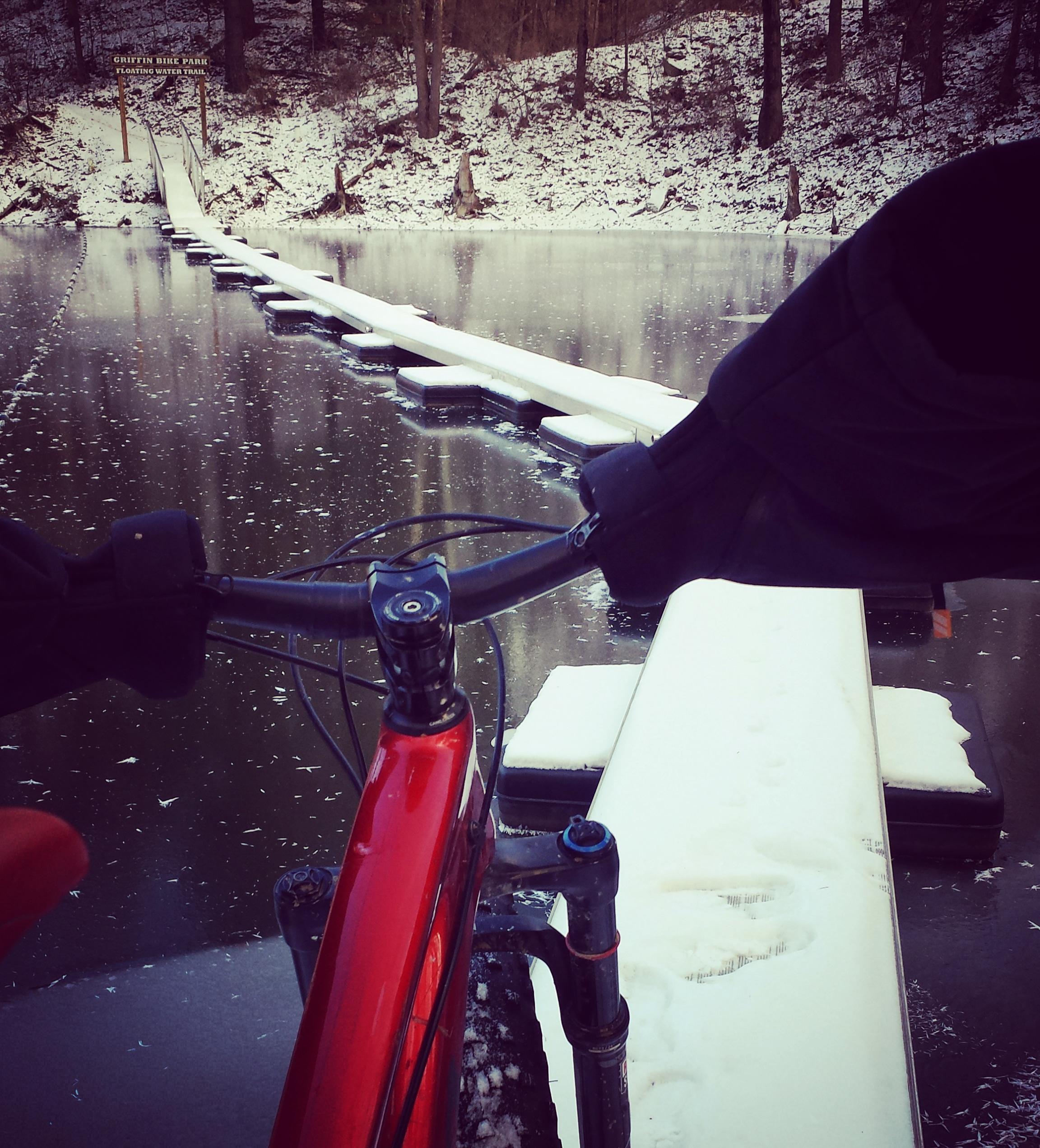A bicycle positioned at the edge of a frozen body of water, with a narrow path of floating platforms leading across. Snow covers the surfaces, and trees are visible in the background. A sign for Griffith Bike Park is partially visible. The scene captures a serene winter landscape. Griffin Bike Park mountain bike trail.