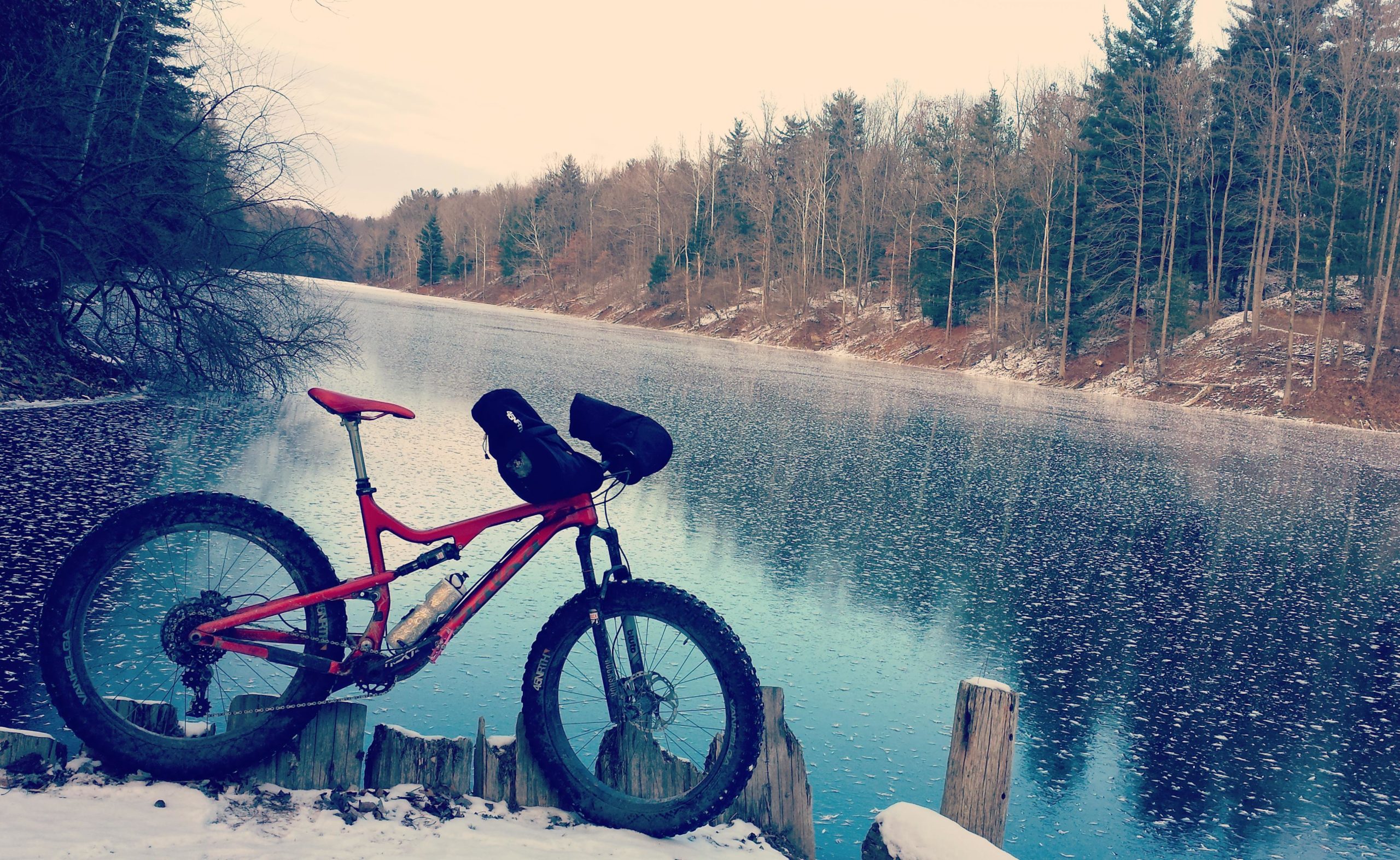 A red mountain bike rests on a wooden post near a calm lake surrounded by trees. The scene features a wintry landscape with snow on the ground and bare branches. The water reflects the trees and the sky, creating a serene atmosphere. Griffin Bike Park mountain bike trail.