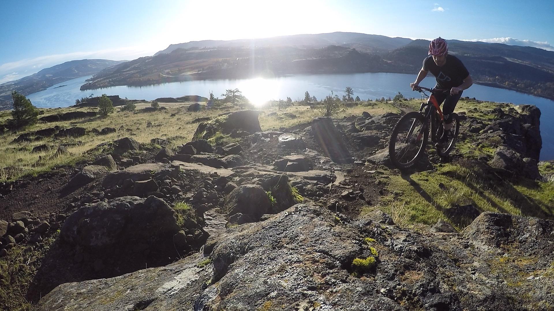 A mountain biker navigating rocky terrain with a scenic river and hills in the background, illuminated by sunlight. Syncline mountain bike trail.